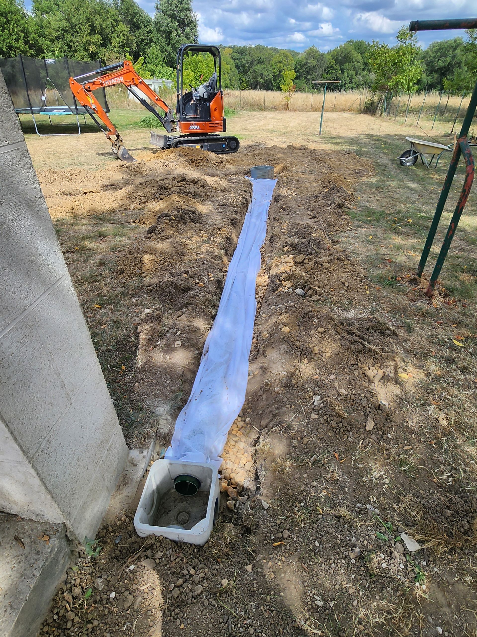 Une petite excavatrice est garée près d'une tranchée revêtue d'une bâche bleue qui mène à un bassin de rétention dans une cour herbeuse.