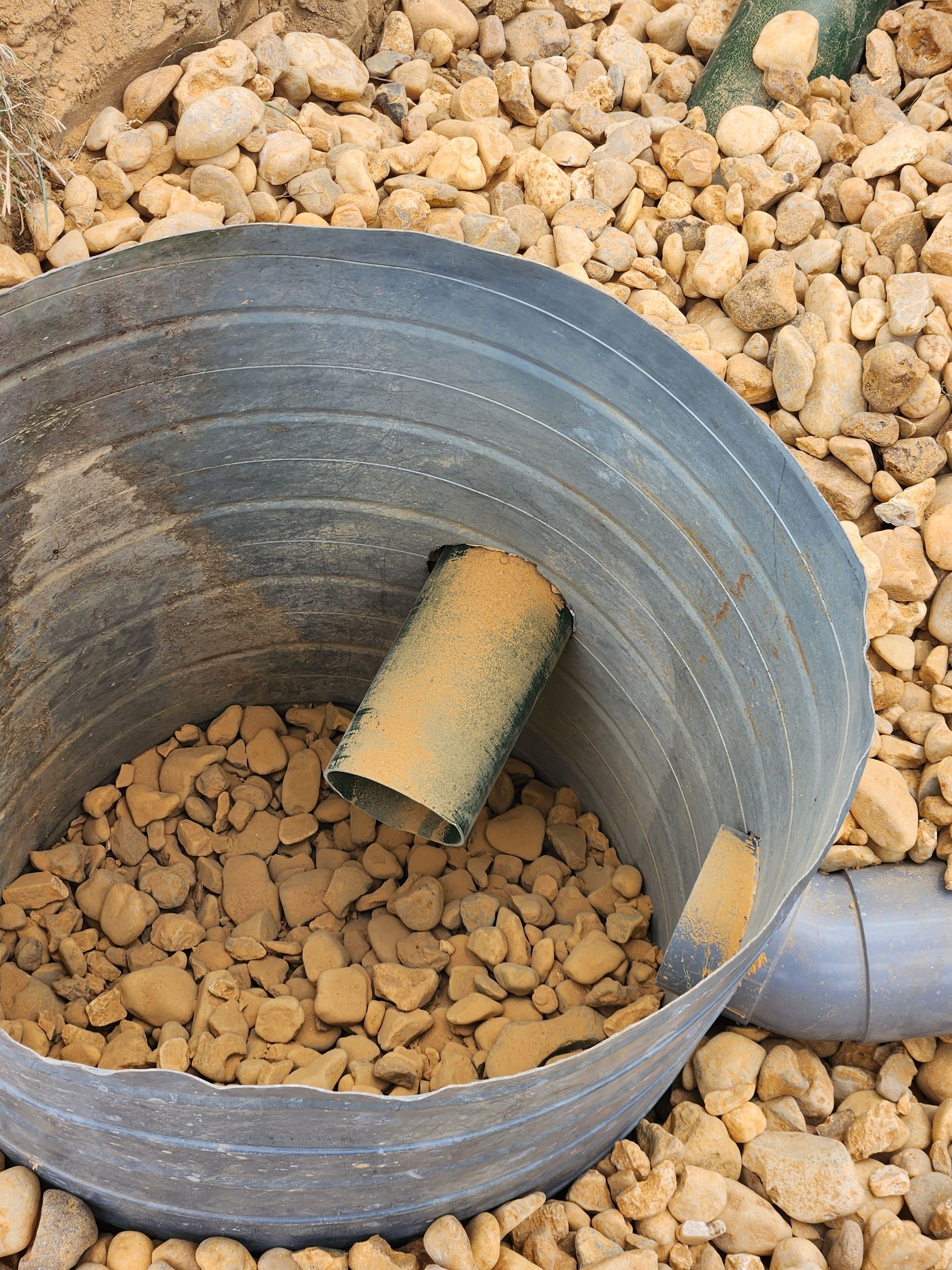 Un bassin de rétention des eaux pluviales en plastique noir ondulé, installé dans du gravier, comportant un tuyau d'arrivée et un coude d'évacuation gris.