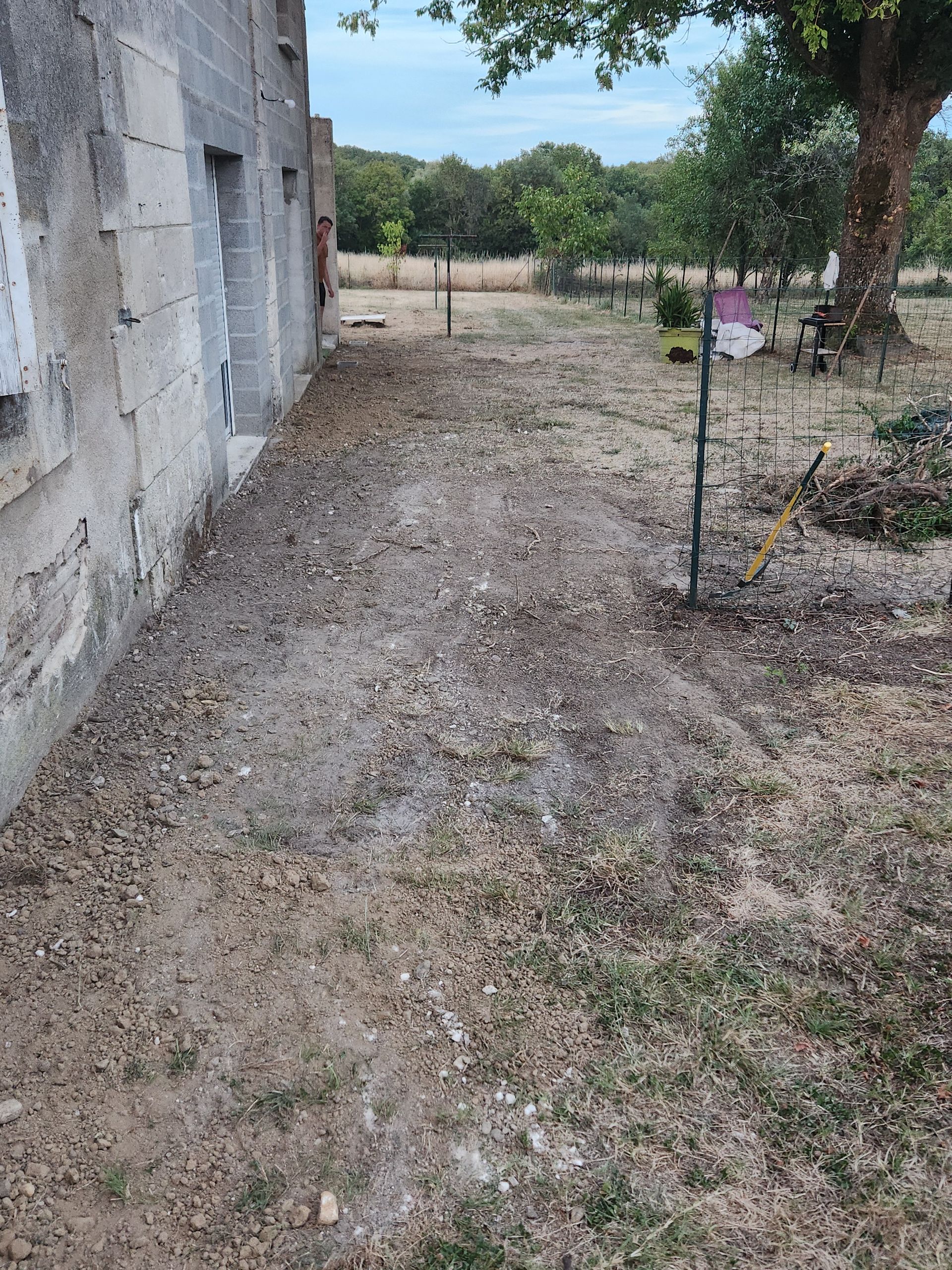 Vue latérale d'une maison en pierre avec une cour partiellement dégagée et recouverte de terre menant à des arbres et à un champ ouvert.