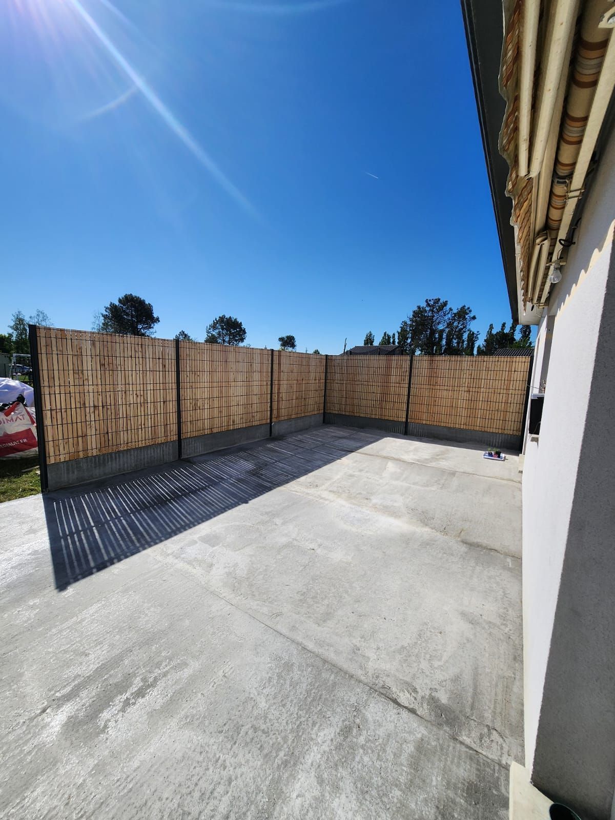 Une terrasse en béton entourée d'une clôture en bambou brun clair, sous un ciel bleu azur.