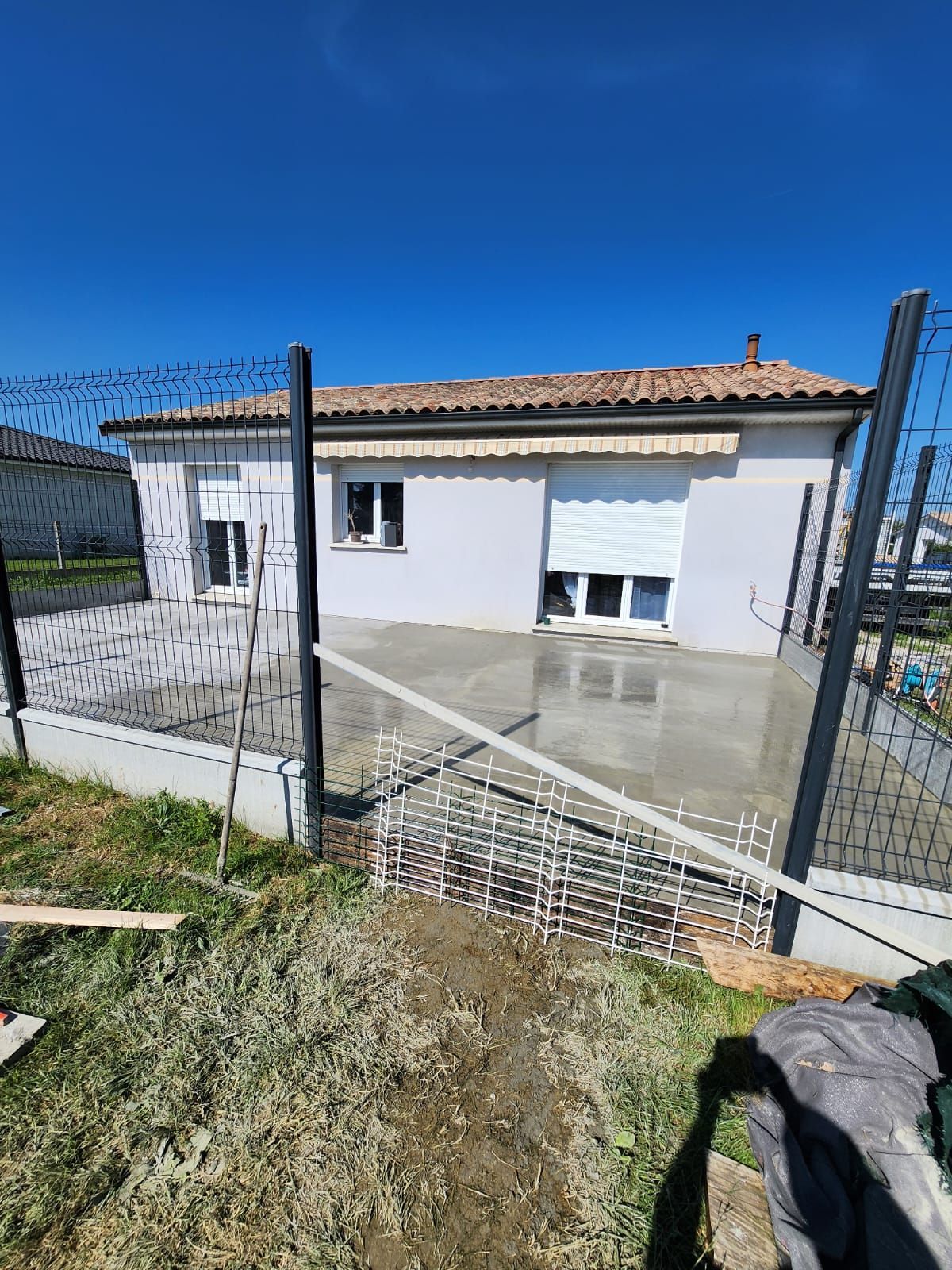 Une terrasse en béton fraîchement coulée se trouve derrière une maison de couleur claire au toit de tuiles, entourée d'une clôture en grillage.