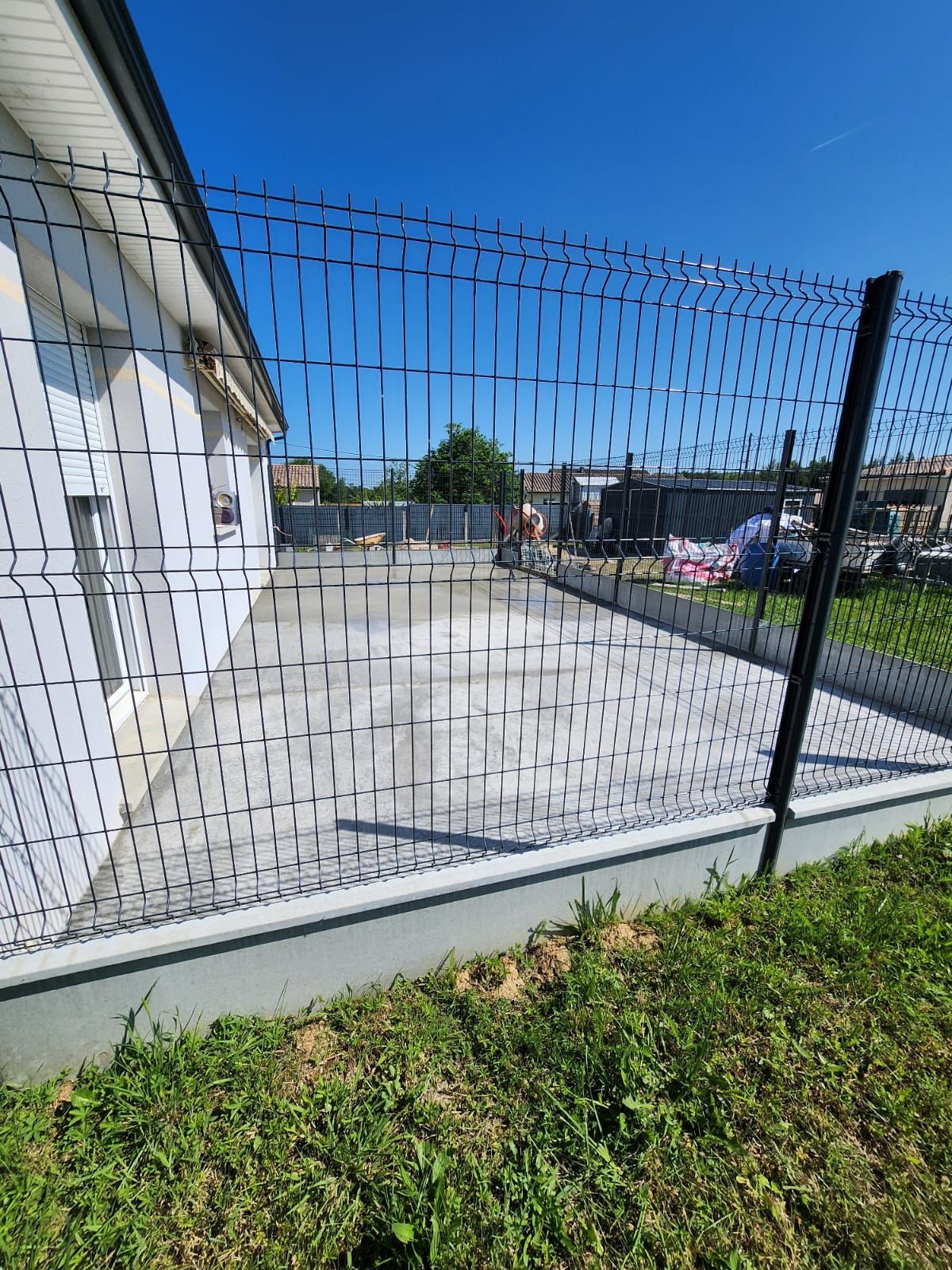 Une terrasse en béton fraîchement coulée jouxte une maison, entourée d'une clôture en métal noir sous un ciel bleu azur.