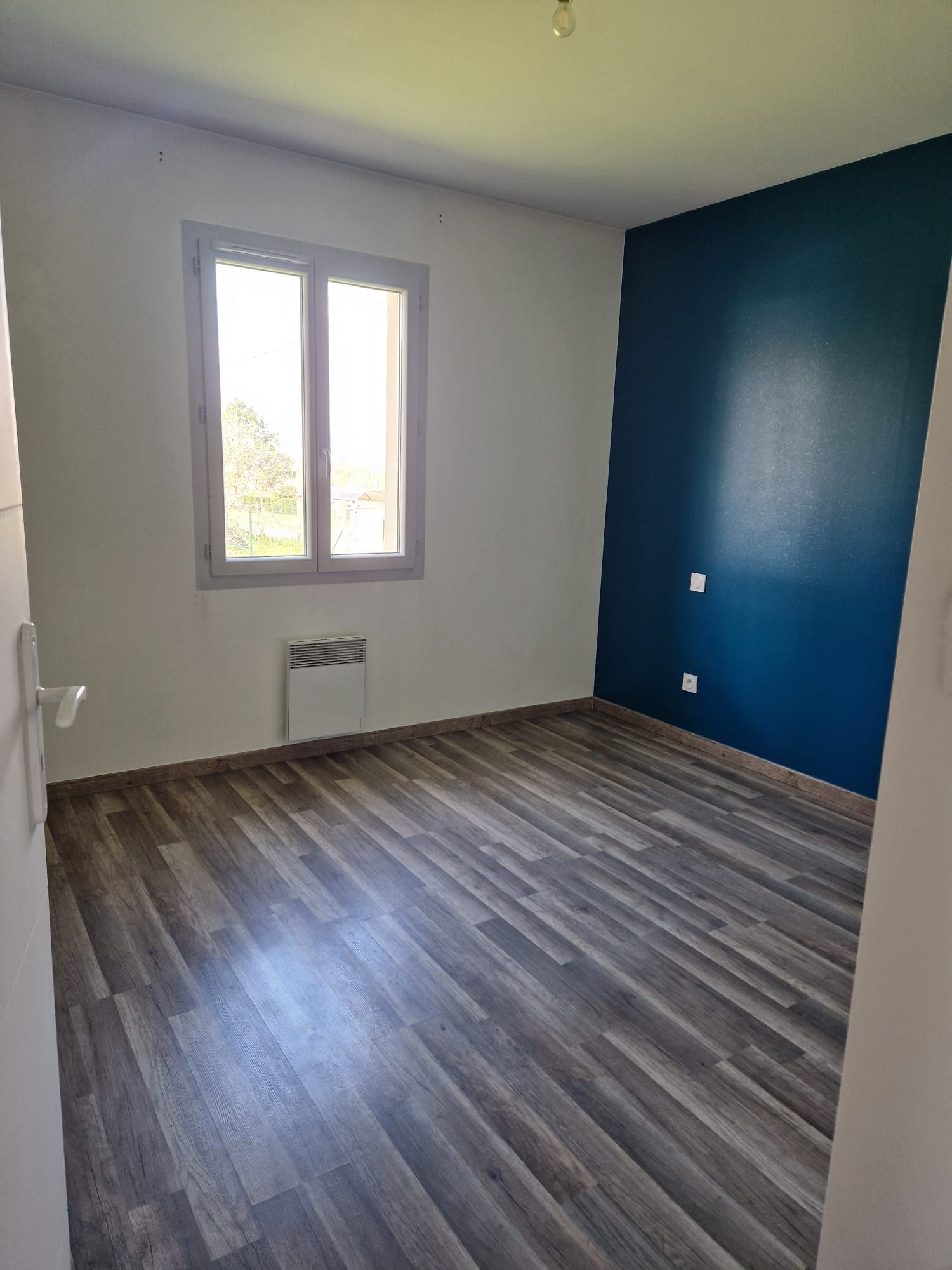 Une chambre vide avec un sol stratifié imitation bois, des murs blancs, un mur d'accent bleu marine, une fenêtre et un radiateur mural.