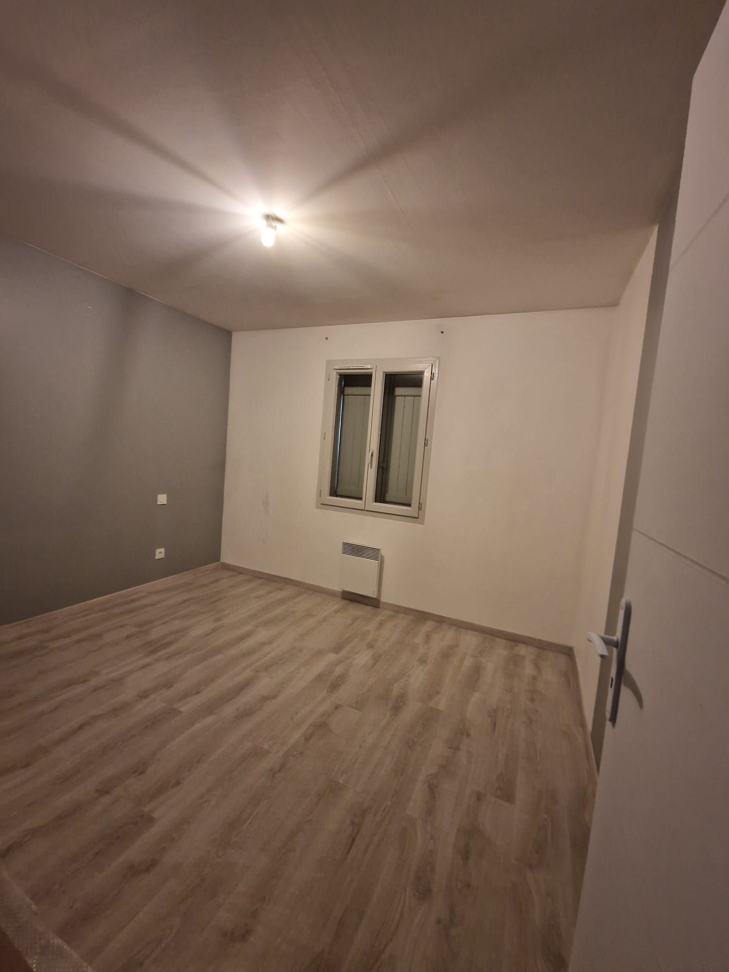 Pièce vide avec un sol clair imitation bois, des murs blancs, un mur d'accent gris foncé, une fenêtre centrale et un plafonnier.