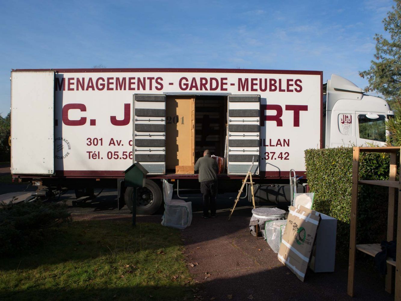 Camion de déménagement portes ouvertes, personne en train de charger. Inscription sur le camion : « MENAGEMENTS - GARDE-MEUBLES CJ », adresse et numéro de téléphone.