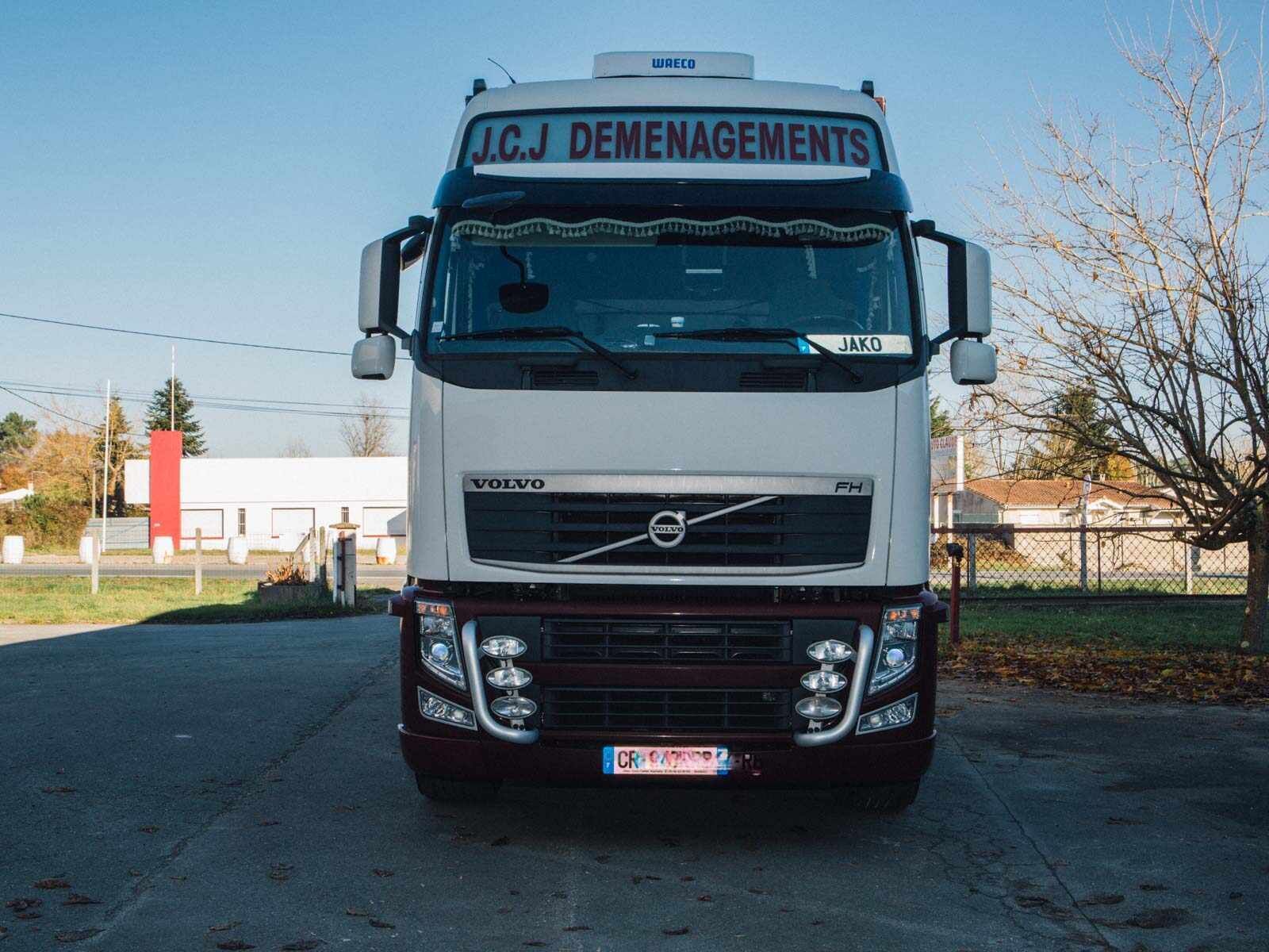 Vue de face d'un semi-remorque Volvo blanc portant l'inscription « JCJ DÉMARCHAGES ». Détails rouge foncé. Stationné sur le trottoir.