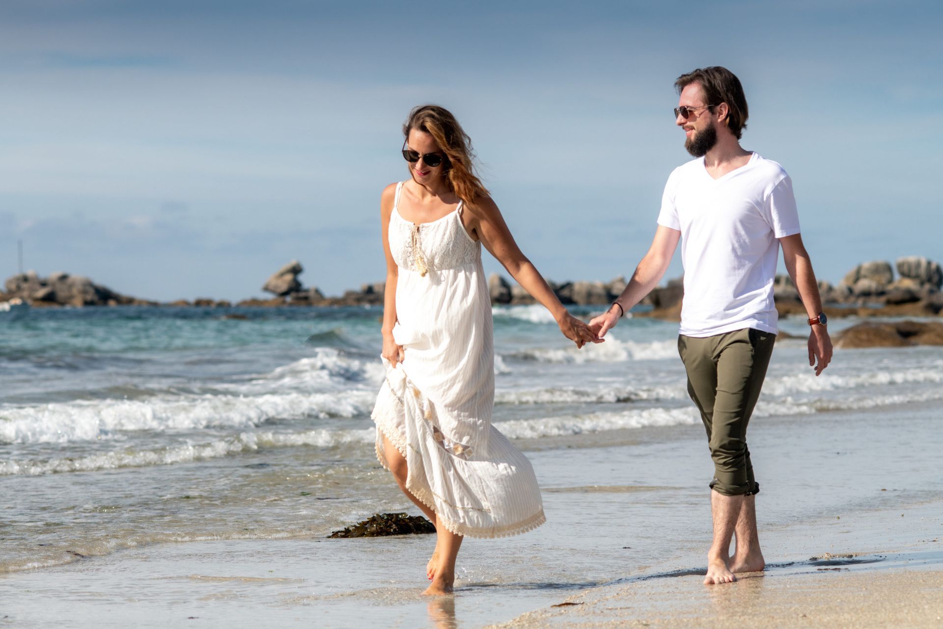 Un couple se tenant la main, marchant pieds nus sur une plage ensoleillée.