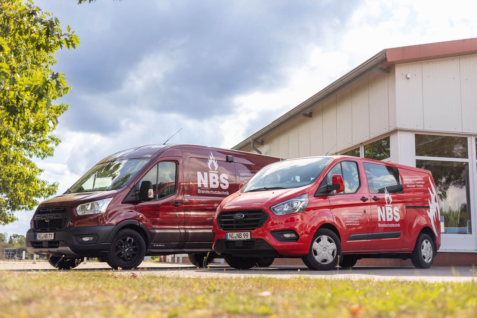 Zwei rote Lieferwagen mit dem „NBS“-Logo parken vor einem Gebäude.