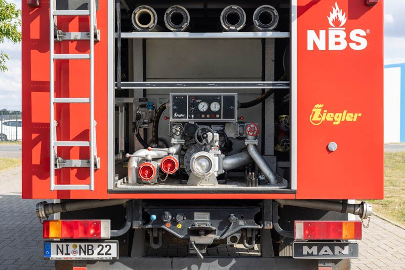 Rückansicht eines roten Feuerwehrautos mit sichtbarer Leiter, Schläuchen und Ausrüstung. „NBS“-Logo und „Ziegler“-Schriftzug vorhanden.