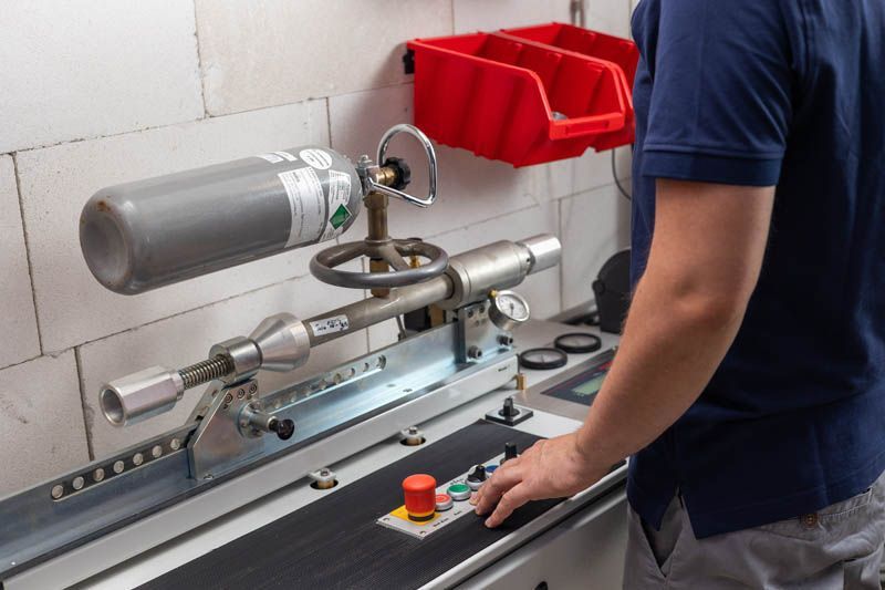 Person, die eine Maschine mit einer grauen Gasflasche bedient. Ein roter Behälter steht an einer weißen Backsteinmauer.