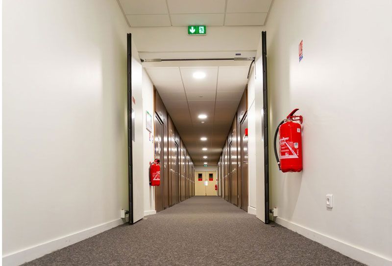 Langer Flur mit Türen, Feuerlöschern und einem Ausgangsschild.