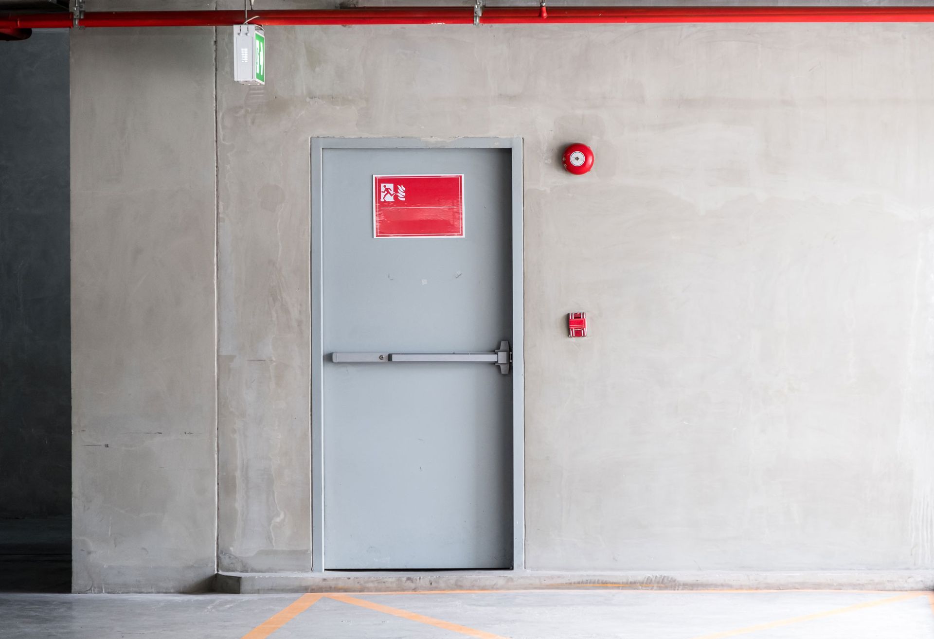 Graue Tür mit rotem Schild, Zugstange, Feueralarm und Ausgangsschild in Betonwand.