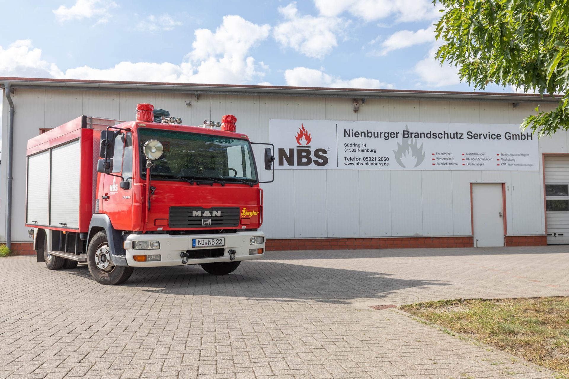 Rotes Feuerwehrauto vor einem weißen Gebäude mit „NBS“-Logo und -Aufschrift geparkt.