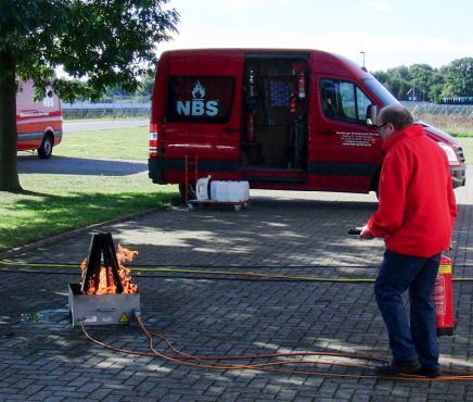 Mann in roter Jacke löscht mit Feuerlöscher brennenden Brand in Metallbox; roter Lieferwagen in der Nähe.
