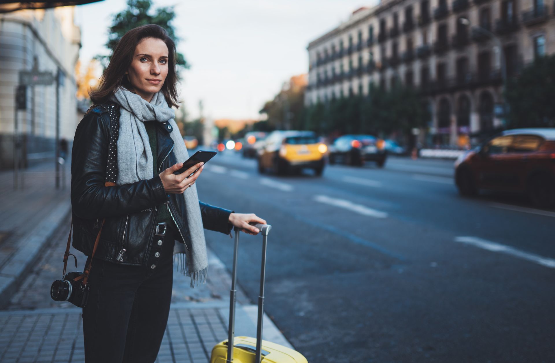 Une femme avec une valise jaune et son téléphone à la main attend un taxi