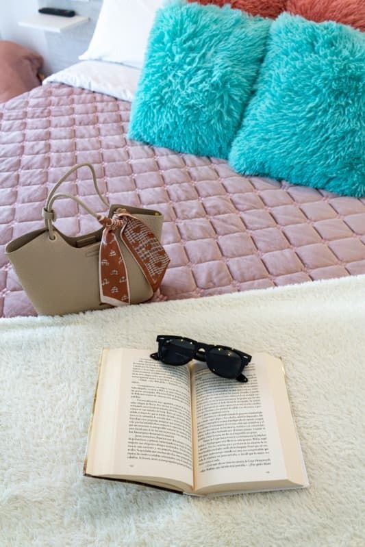 Un libro y unas gafas de sol están sobre una cama al lado de un bolso.