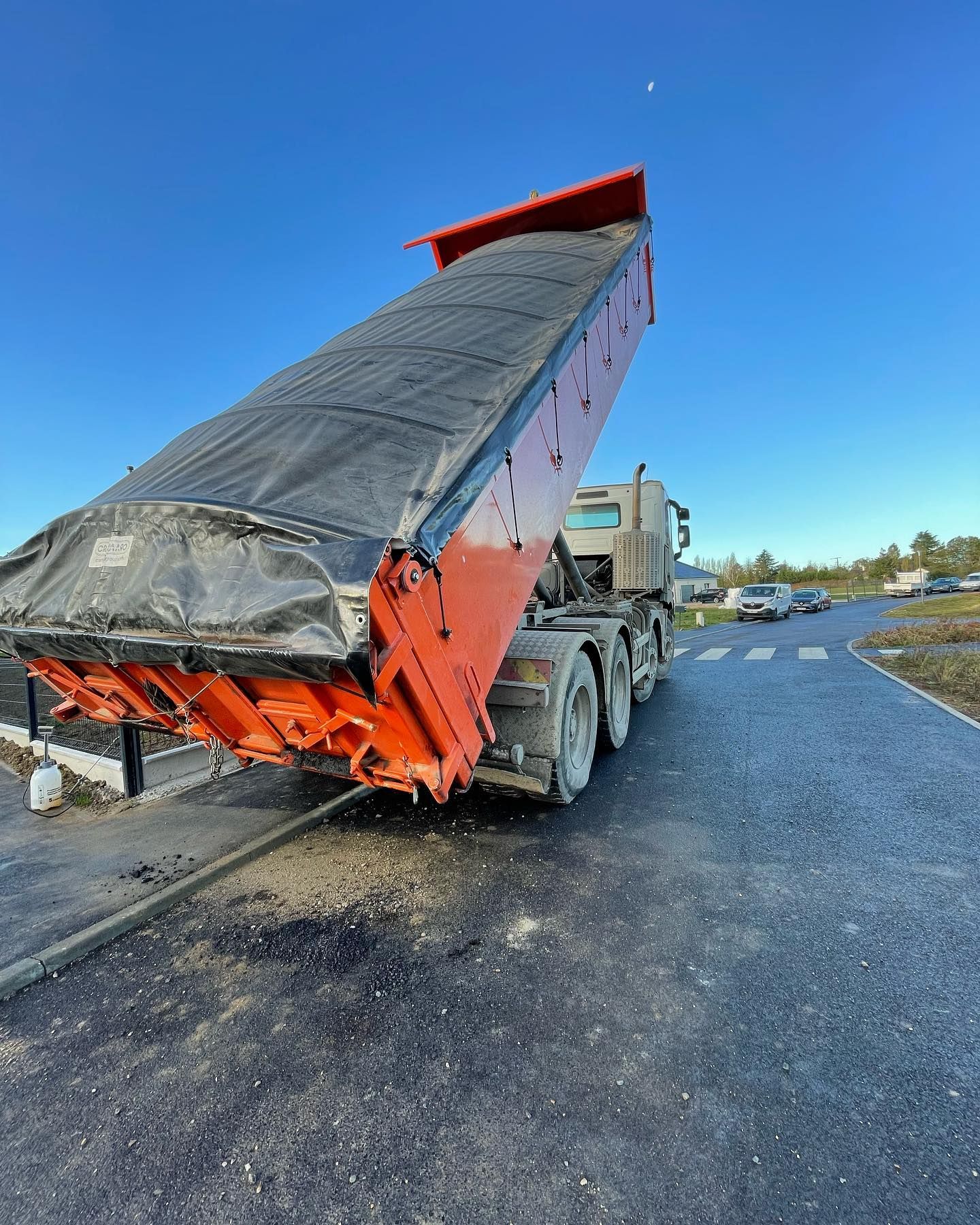 Camion-benne à plateau surélevé déchargeant du gravier sur une route goudronnée ; plateau orange vif, ciel bleu.