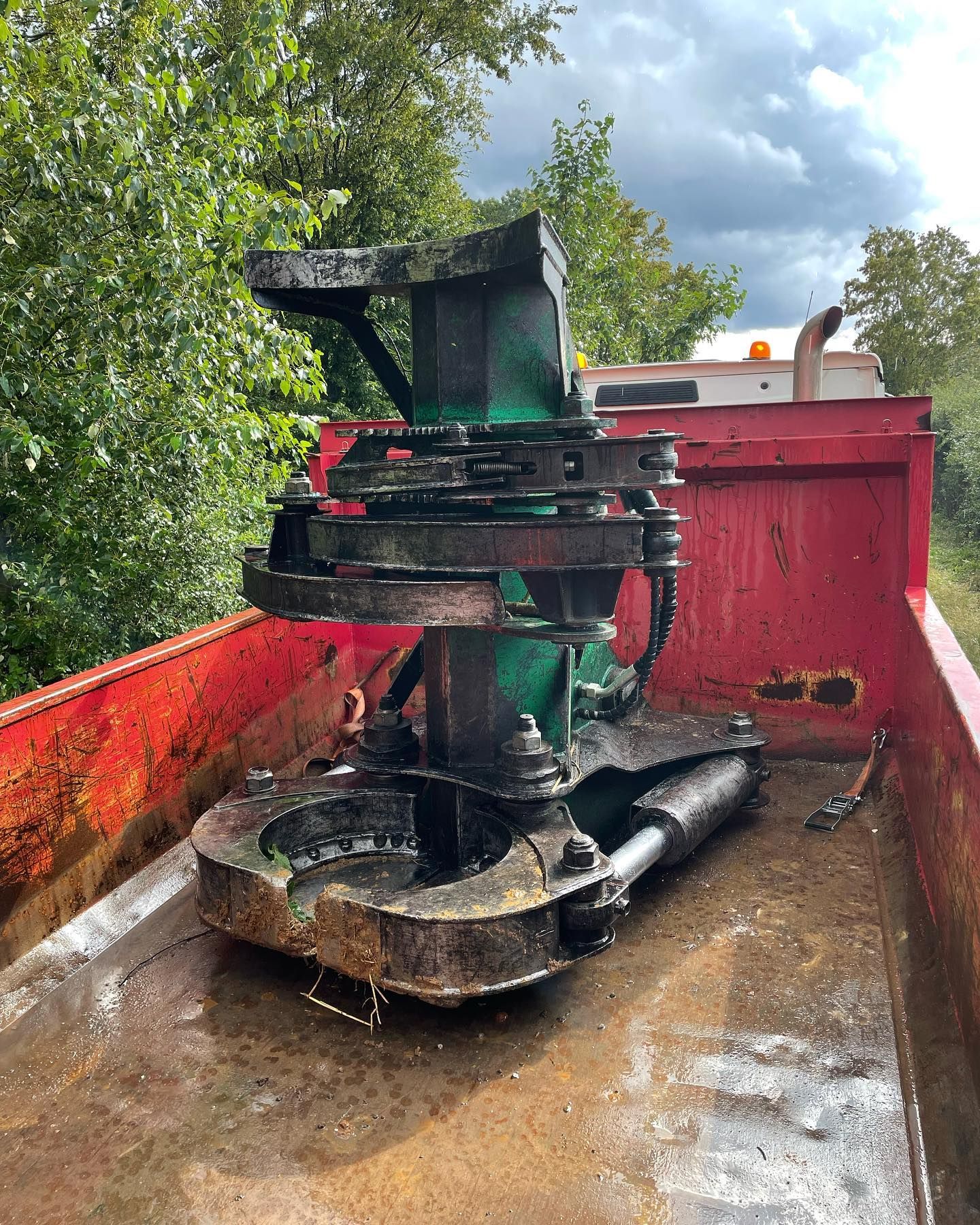 Une tête d'abattage industrielle, sale et usée, est posée à l'extérieur, dans la benne rouge d'un camion vert et noir rouillé.