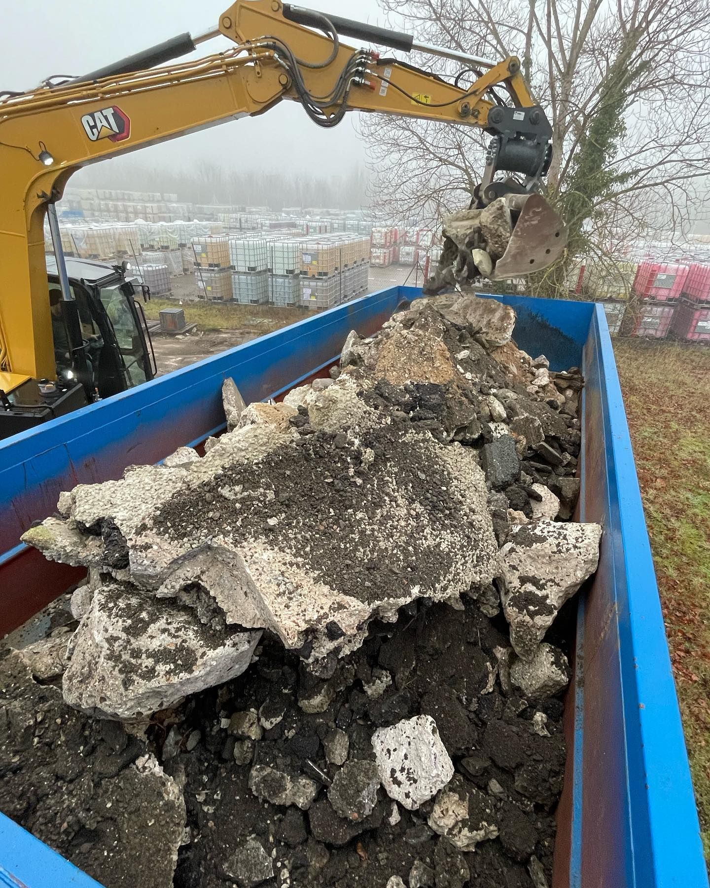 Une pelleteuse jaune charge des débris dans un conteneur bleu à l'extérieur.