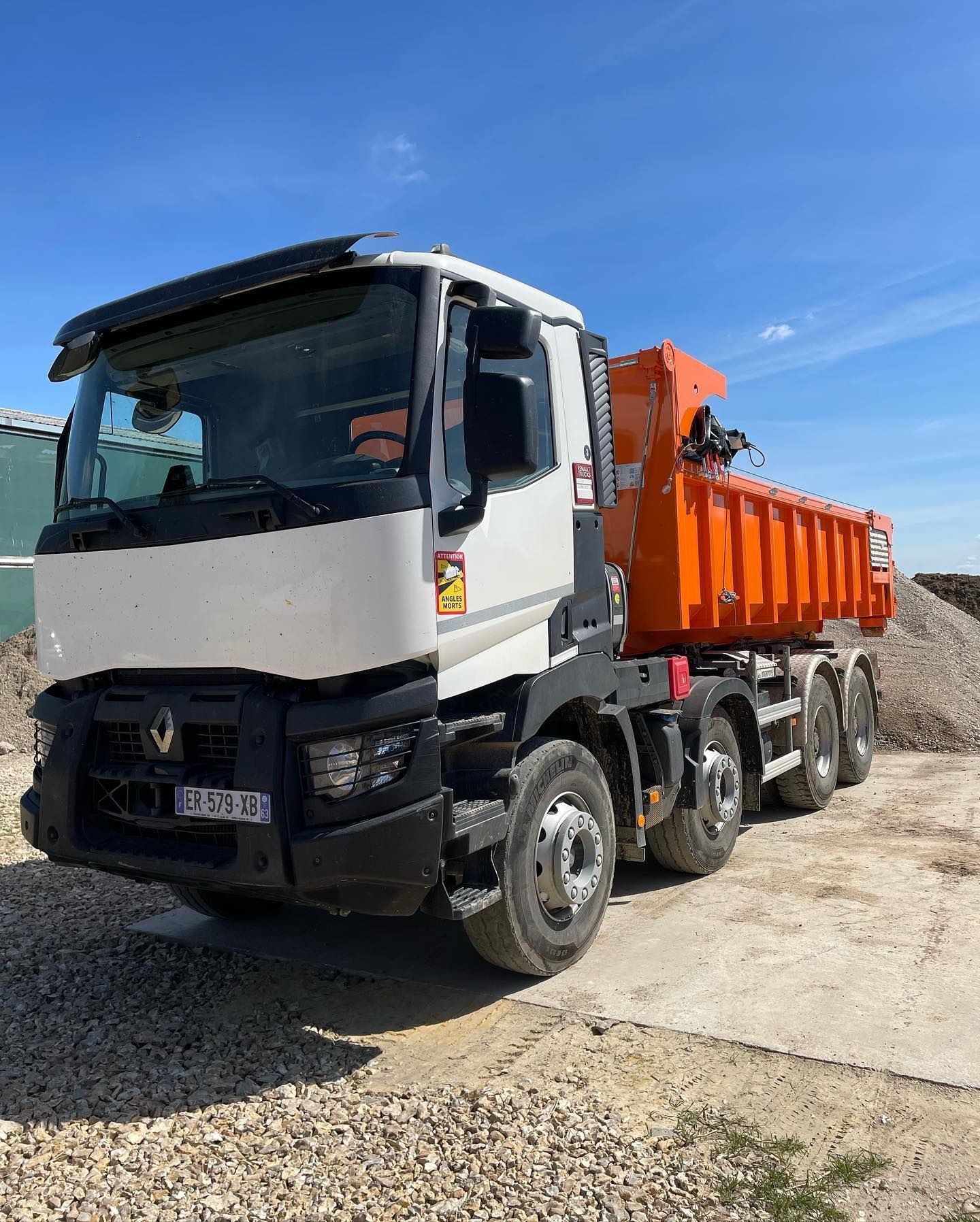 Camion-benne blanc et noir à benne orange, stationné sur du gravier. Ciel bleu en arrière-plan.