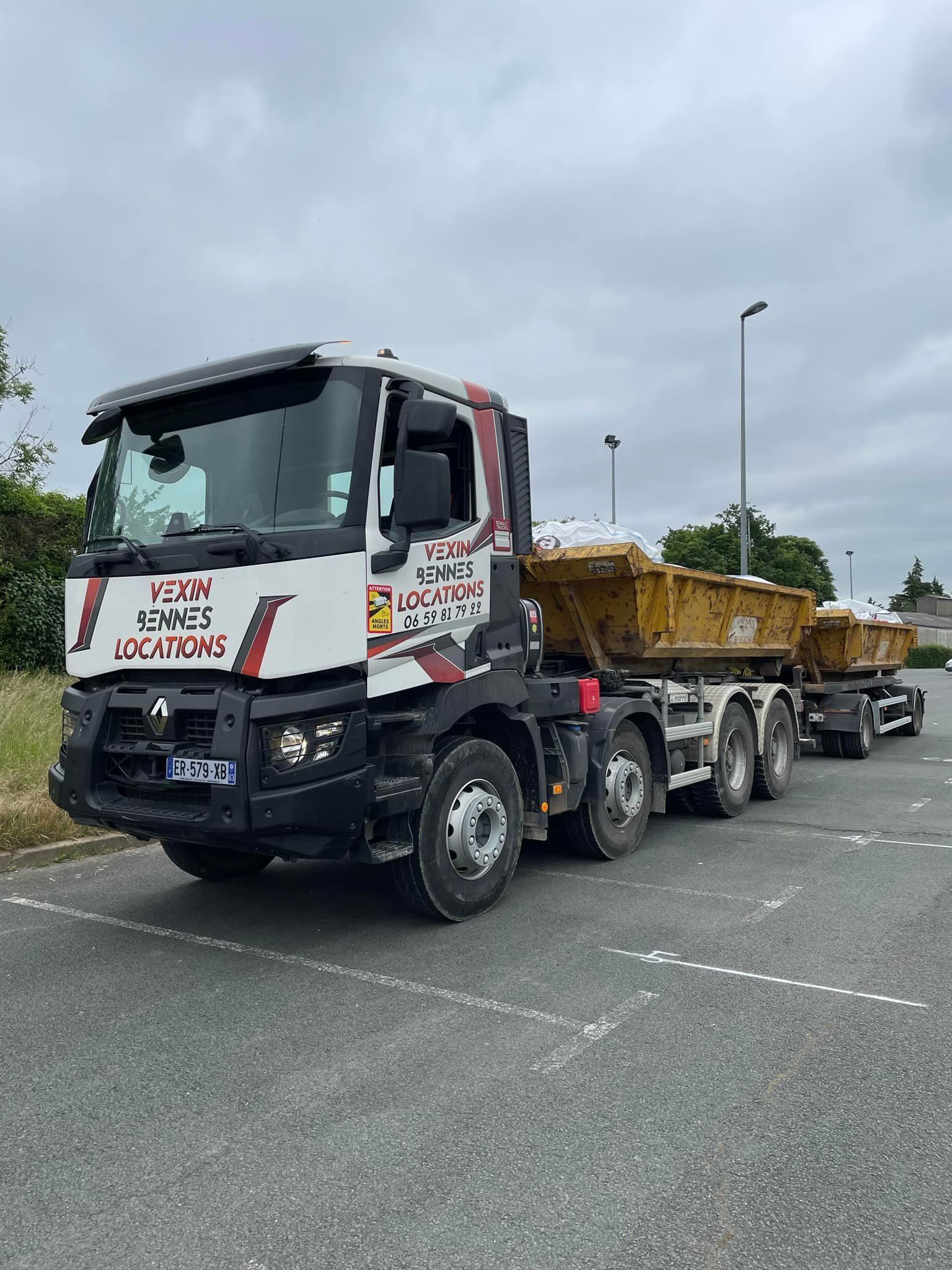 Un semi-remorque blanc et rouge transportant deux bennes basculantes jaunes est stationné sur un parking.