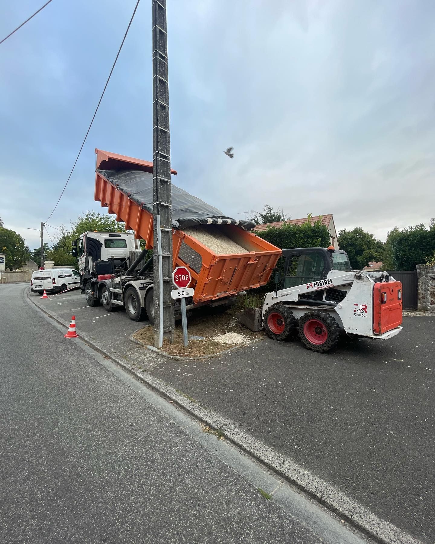 Un camion-benne décharge du sable au sol près d'un poteau électrique, à côté d'une chargeuse compacte.