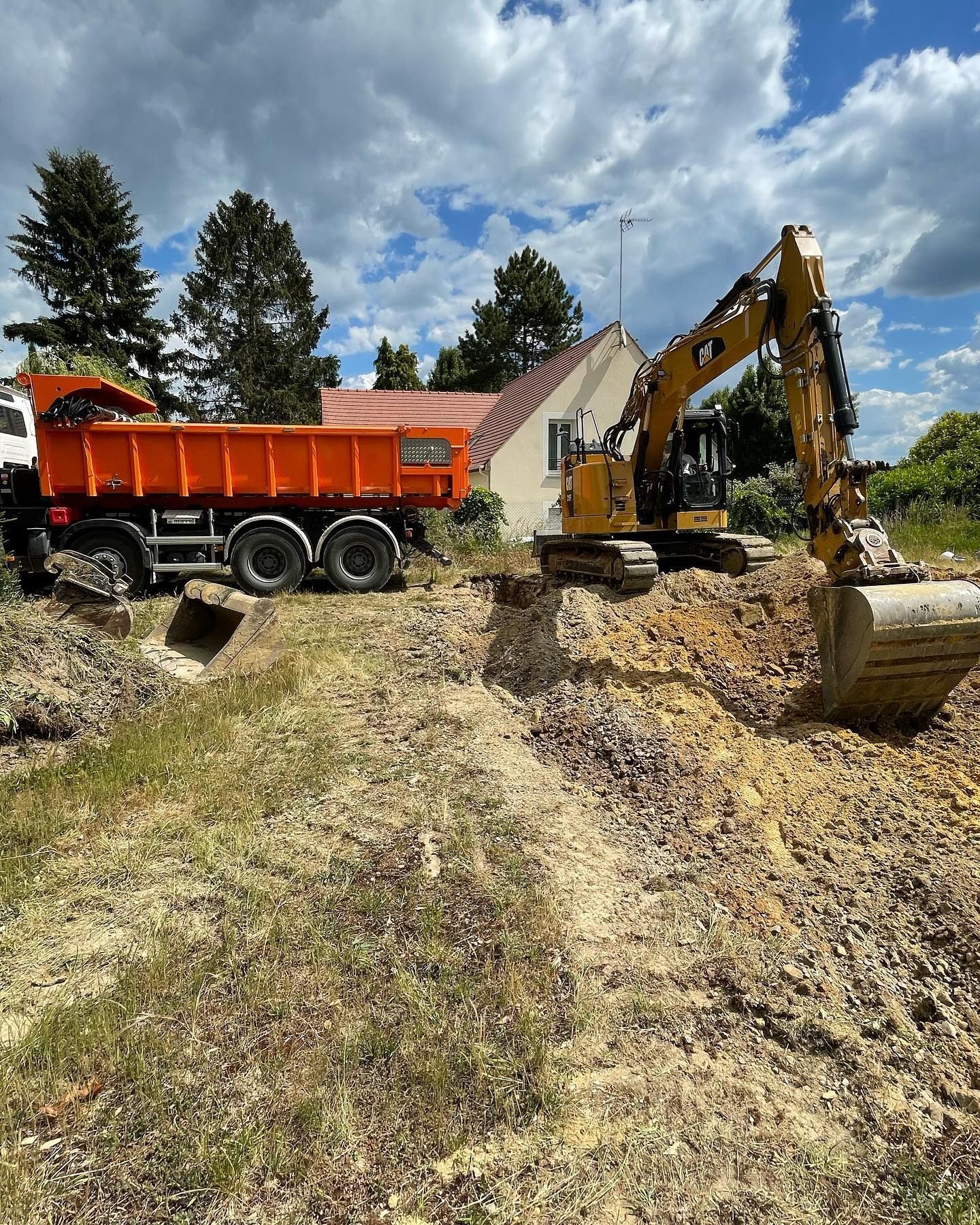 Une excavatrice charge de la terre dans un camion-benne orange sur un chantier de construction près d'une maison, sous un ciel nuageux.