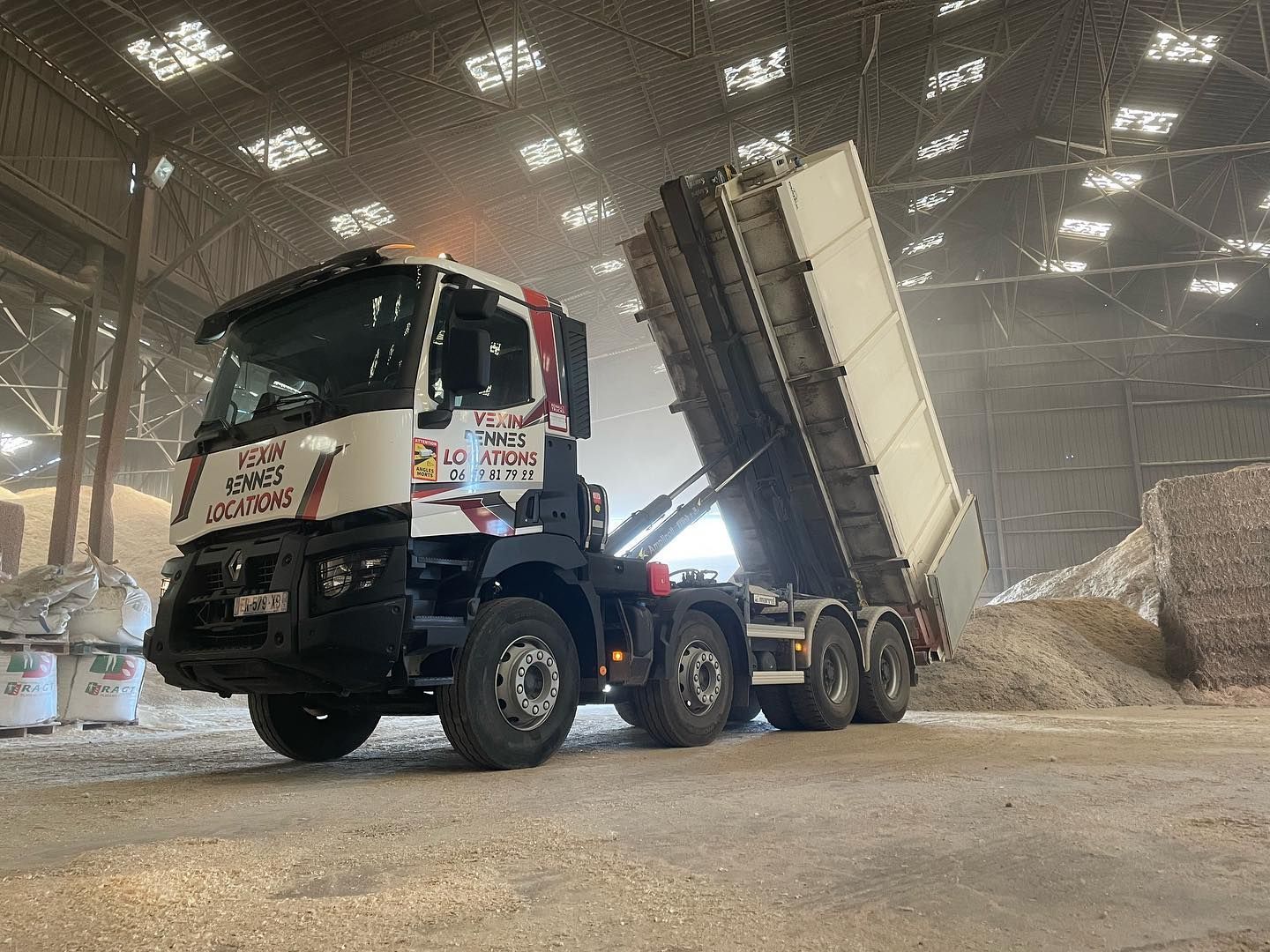 Un camion-benne décharge des matériaux à l'intérieur d'un entrepôt. Le camion est blanc et rouge.