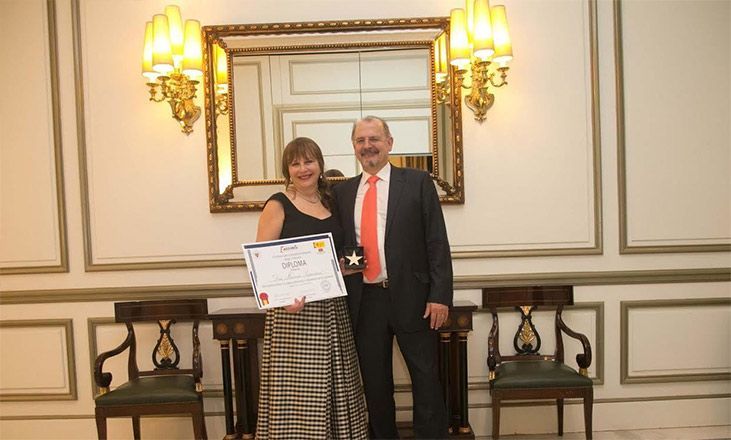 Dos personas posando con un certificado en una elegante habitación con un gran espejo y apliques de pared.