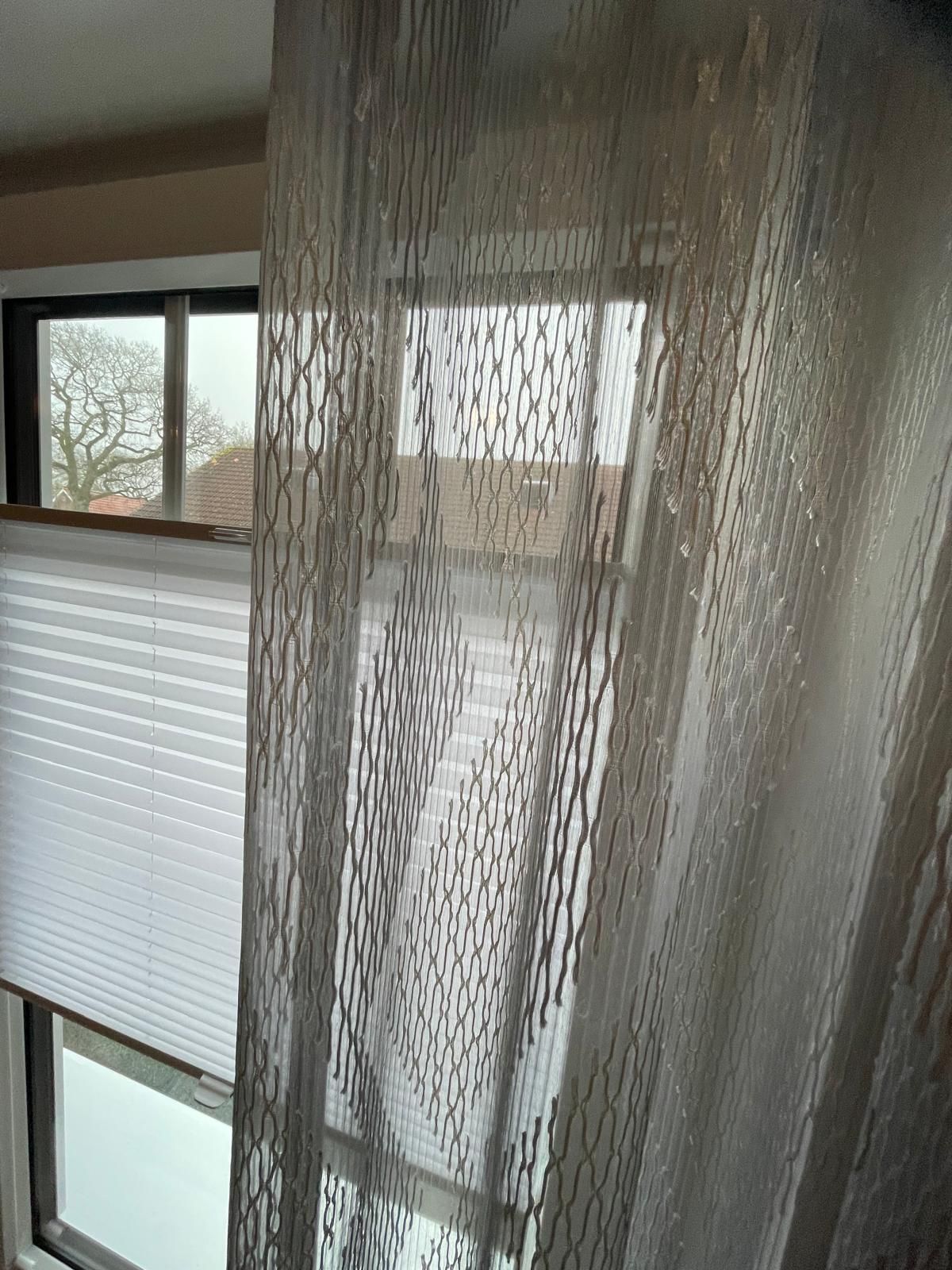 An einem Fenster neben einem Fenster mit Jalousien hängt ein weißer Vorhang.