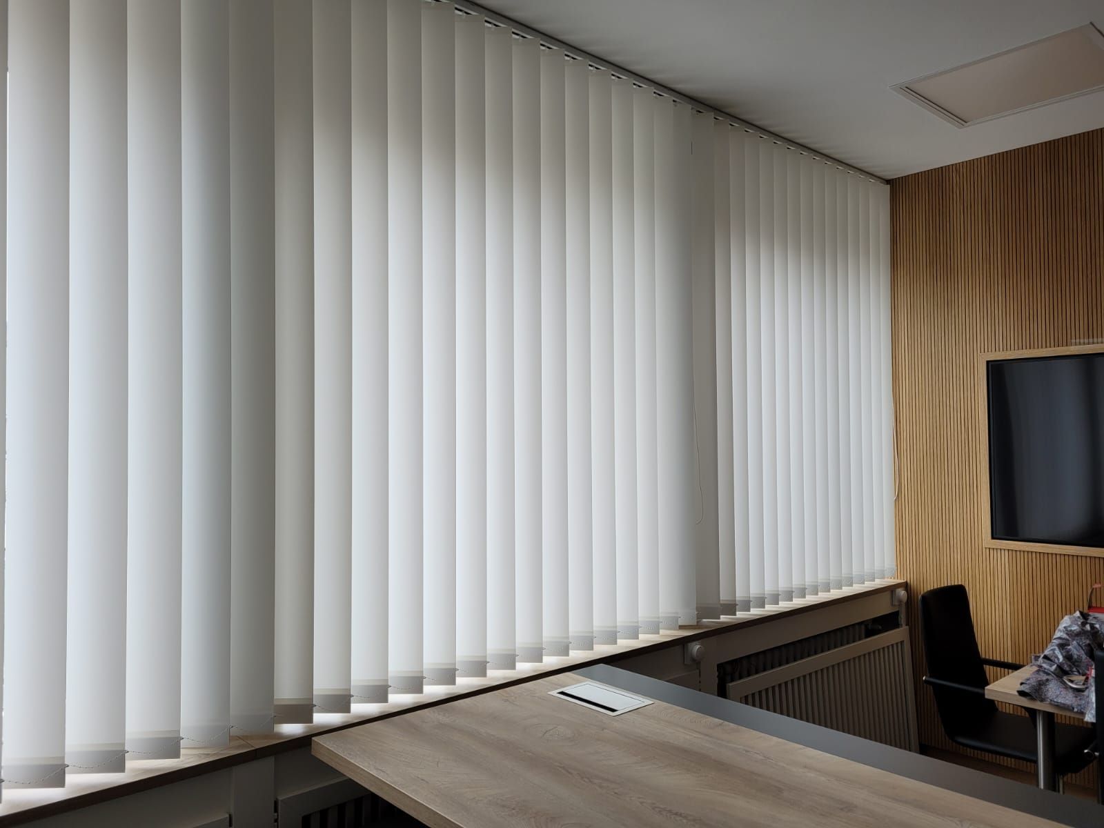 Ein Raum mit weißen Vertikaljalousien an den Fenstern und einem Holztisch.