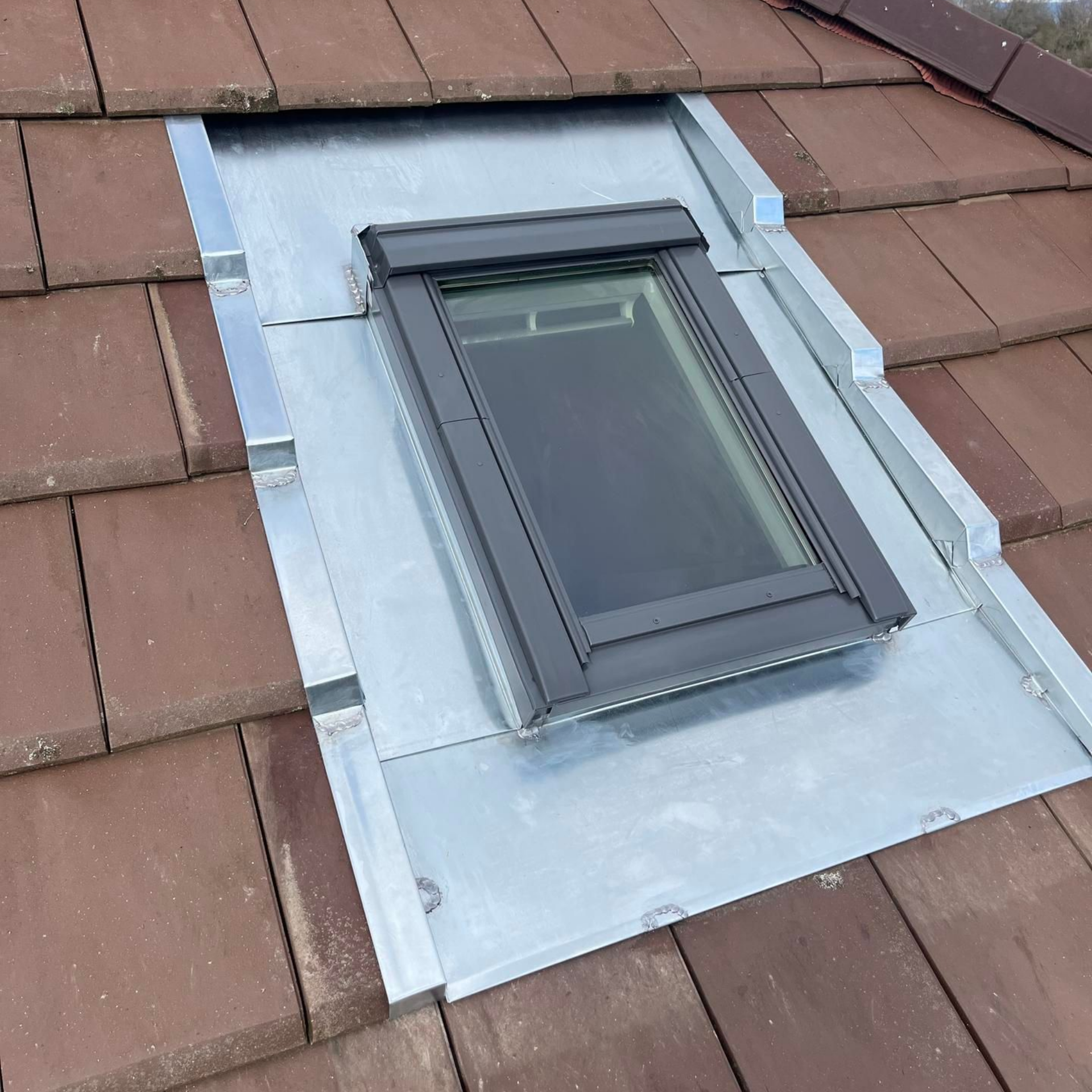 Puits de lumière sur un toit en tuiles brunes, entouré de solins argentés.