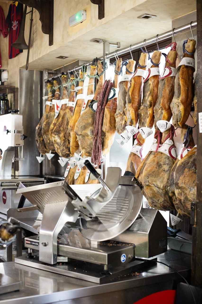 Jamones curados en lonchas colgando en una charcutería, con una cortadora de carne sobre el mostrador.