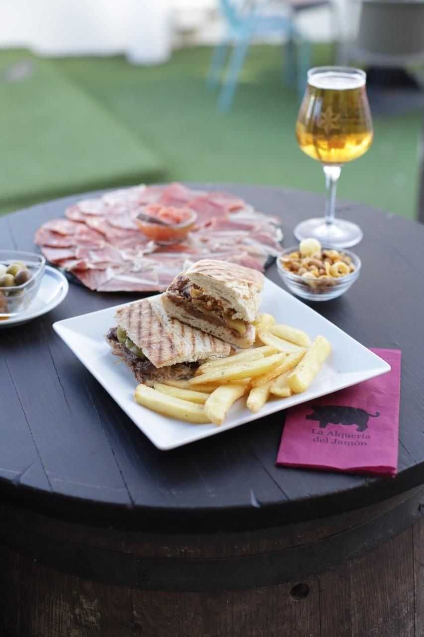 Mesa al aire libre con un sándwich a la parrilla, papas fritas, carne en rodajas y un vaso de cerveza en un patio.