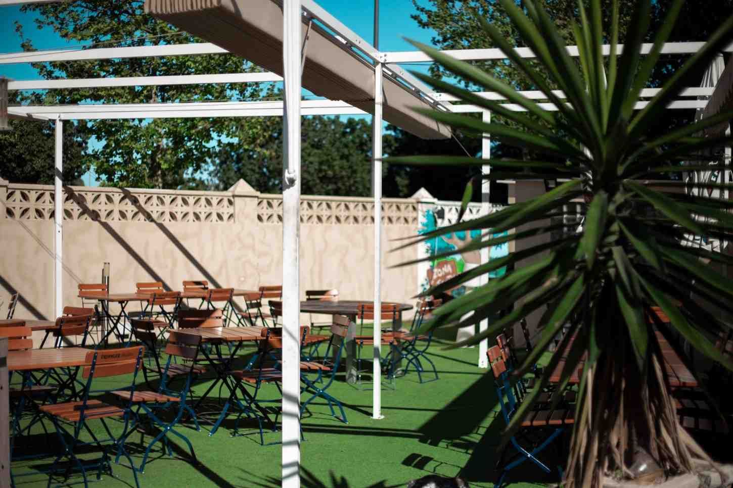 Terraza de cafetería al aire libre con mesas, sillas y una gran planta en maceta bajo vigas blancas de pérgola.