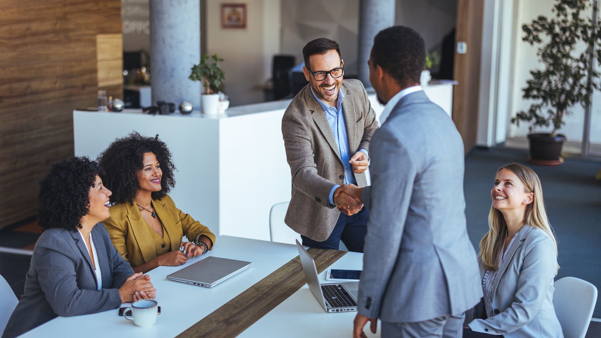 Des hommes d'affaires se serrent la main dans un bureau lumineux. Des collègues souriants sont assis à une table.