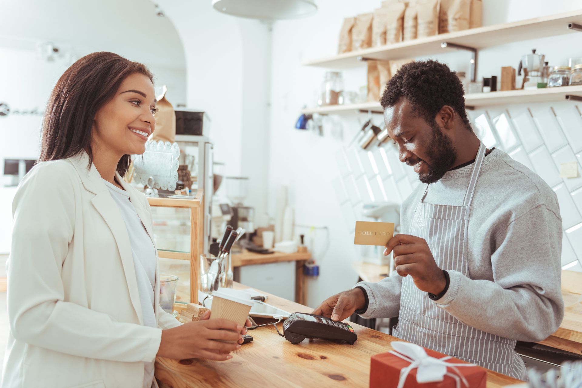 Une femme paie par carte au comptoir d'un café ; le barista tient la carte près du terminal de paiement.