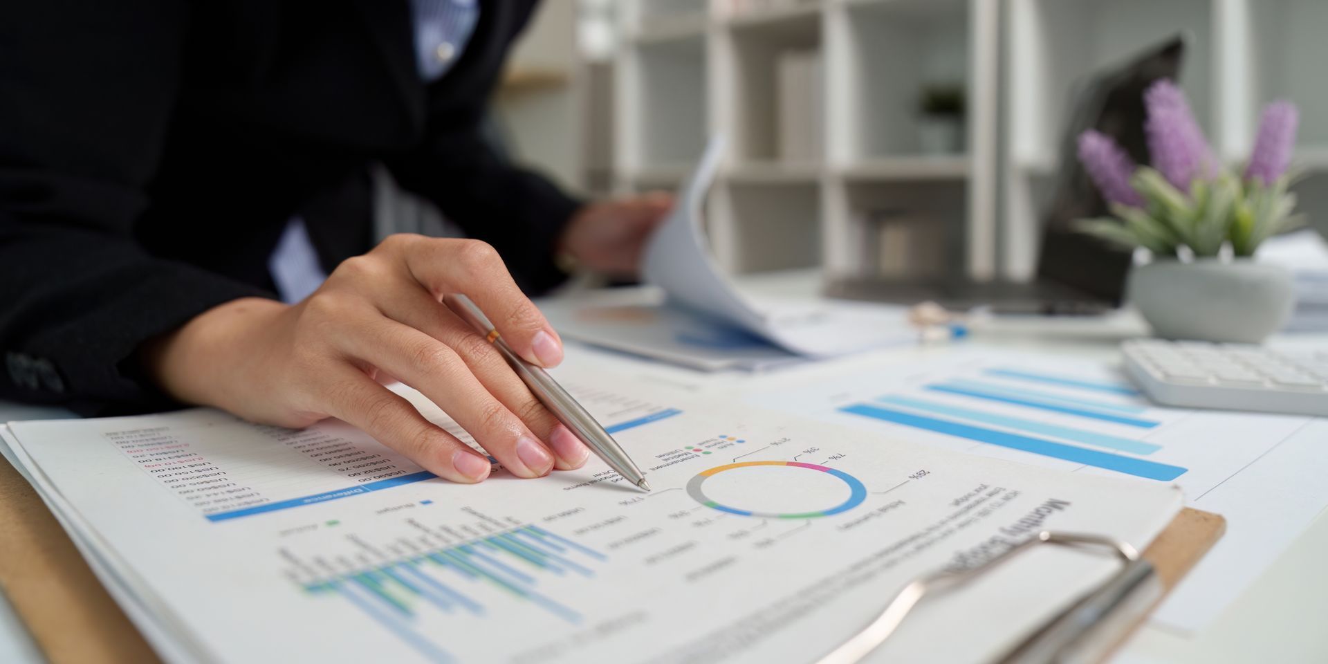 Une personne en blazer désigne des graphiques financiers au stylo. On aperçoit des papiers, un bureau et une plante en pot en arrière-plan.