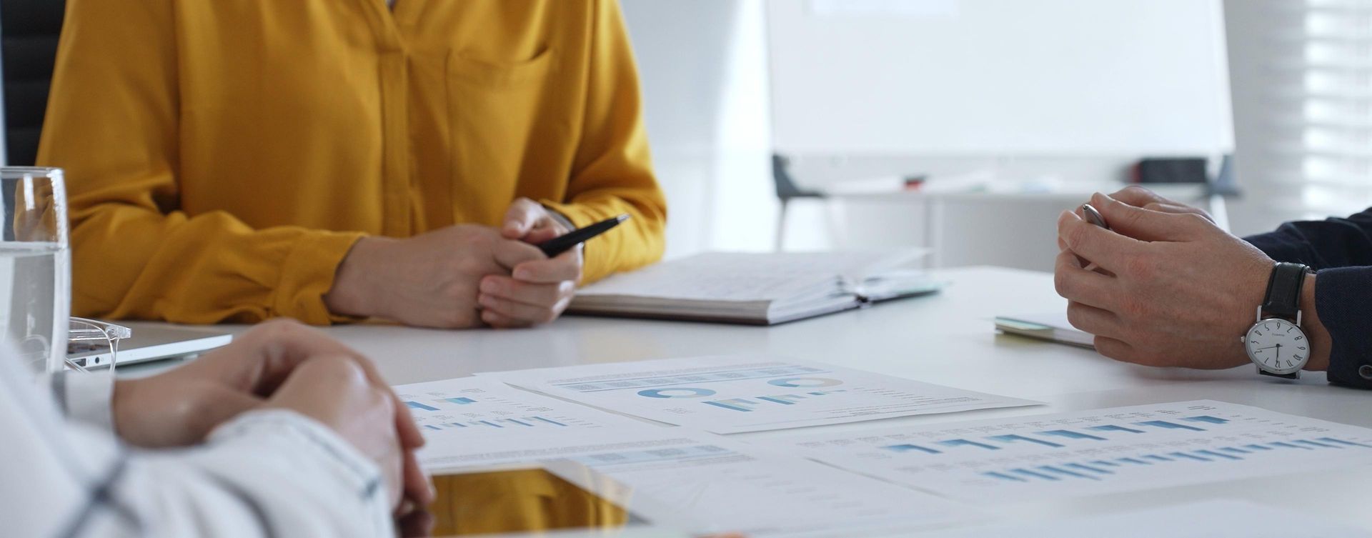 Dans un bureau, trois personnes sont assises à une table blanche. Elles tiennent des stylos et consultent des documents.