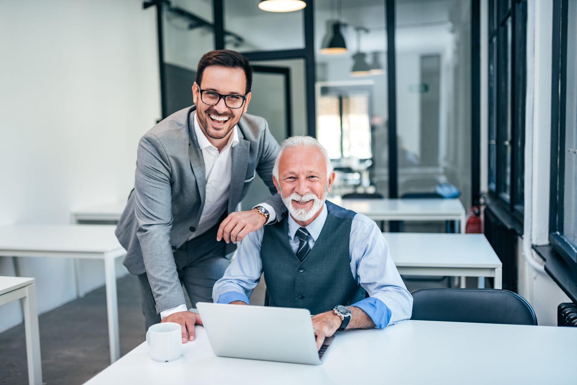 Deux hommes sourient à la caméra dans un bureau ; l'un est appuyé sur l'autre, assis à un bureau avec un ordinateur portable.