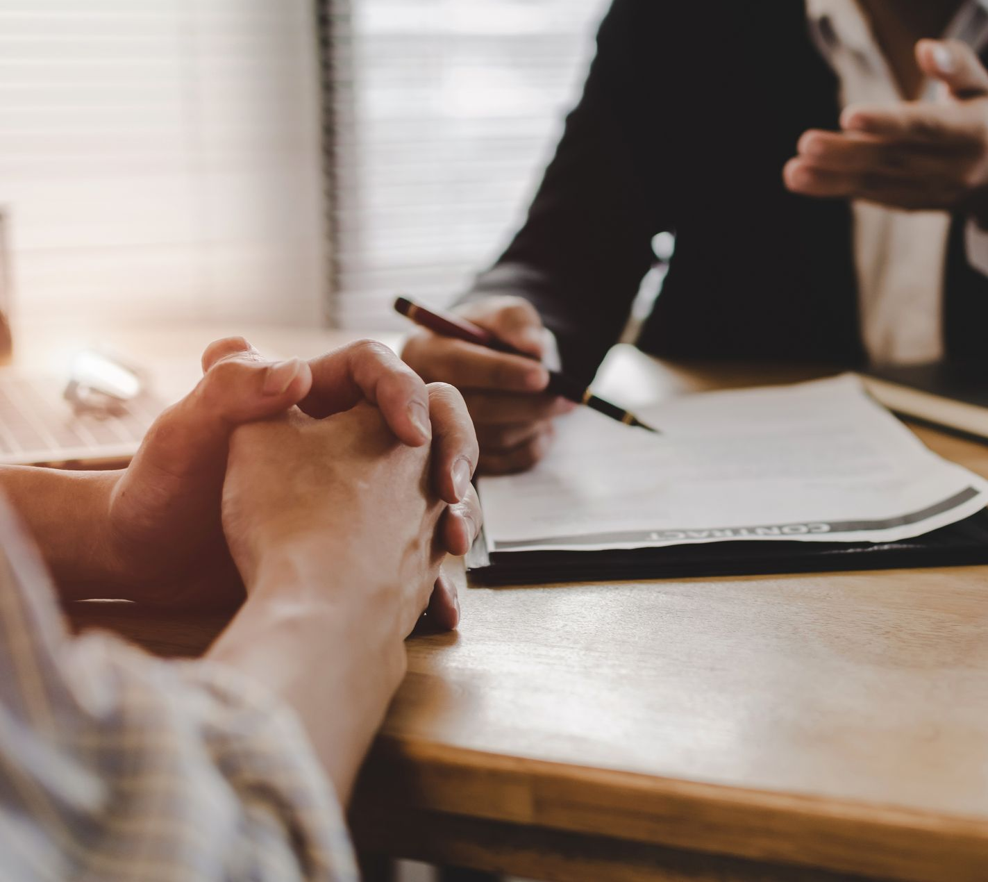 Une personne, les mains jointes, est assise à une table face à une personne tenant un stylo au-dessus de papiers.