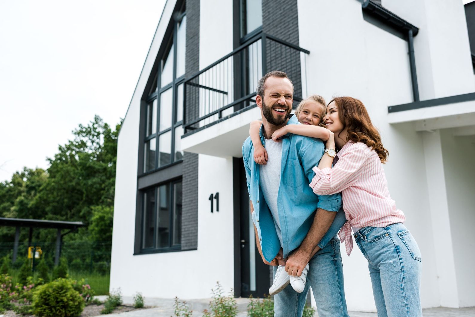 Une famille devant une maison blanche moderne ; le père porte sa fille sur son dos, la mère sourit.