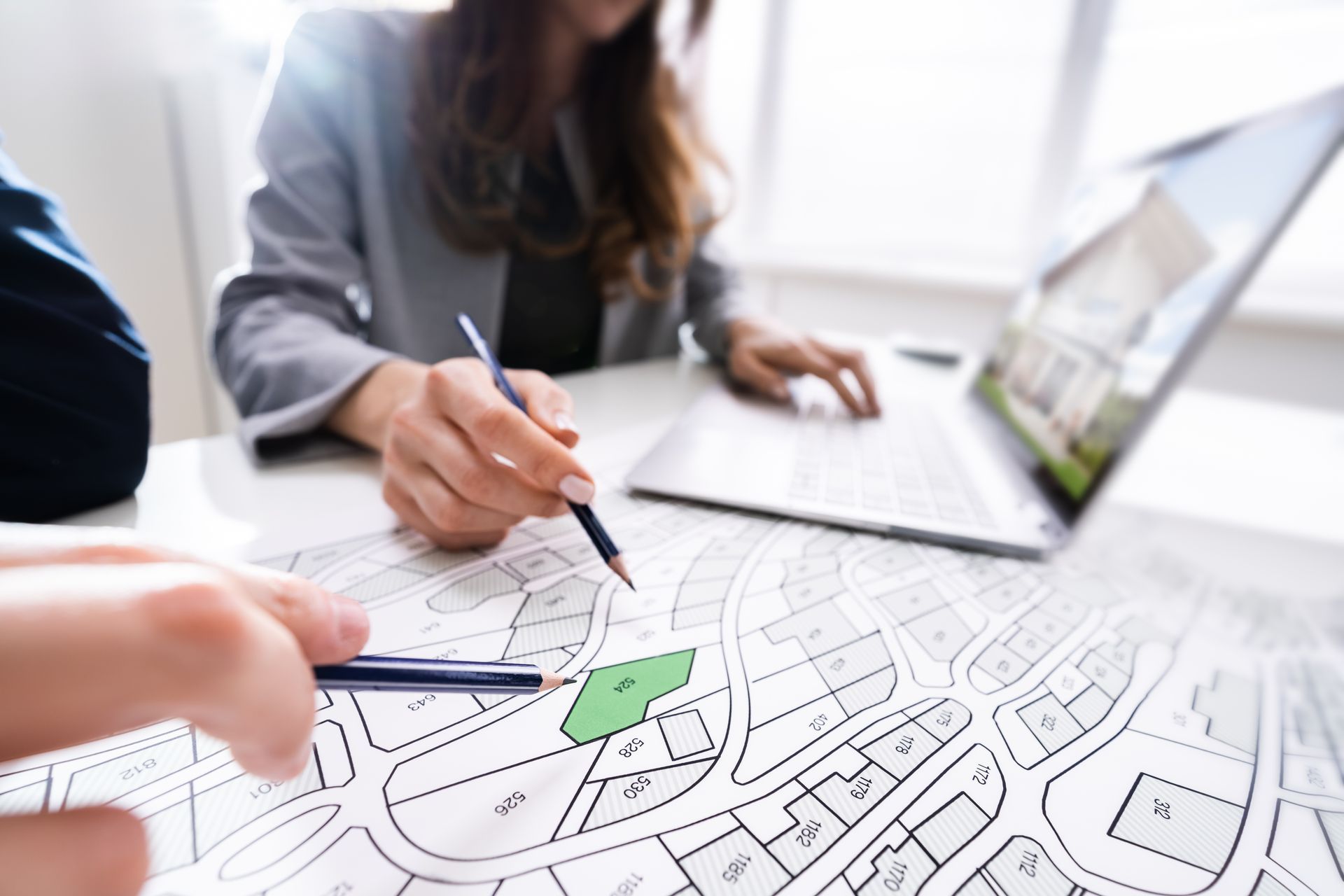 Deux personnes consultent une carte topographique, l'une pointant du doigt avec un stylo, l'autre utilisant un ordinateur portable affichant l'image d'une maison, à l'intérieur.