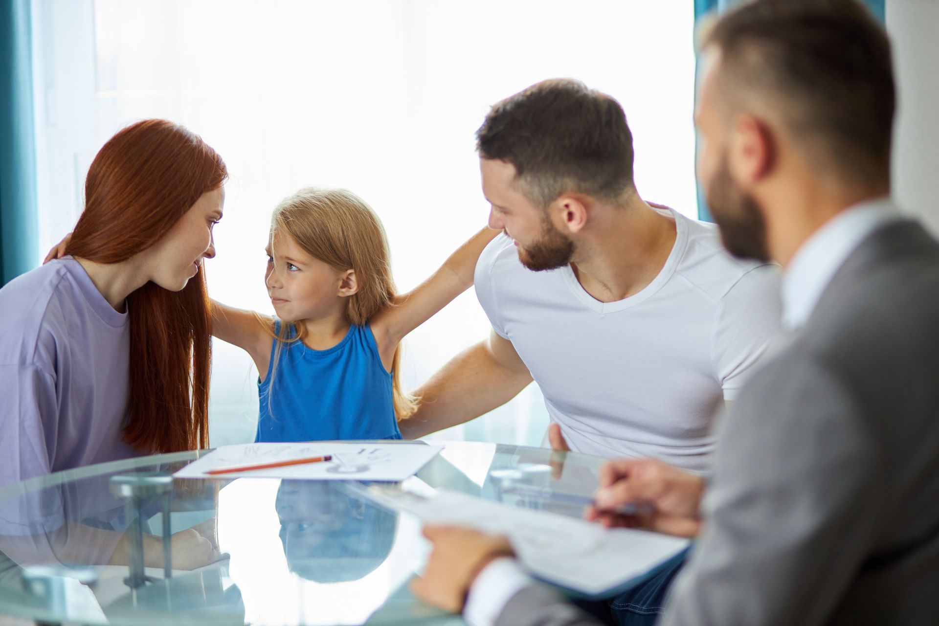 Une famille est assise à une table en verre avec un professionnel ; l'enfant fait des gestes en regardant un parent.