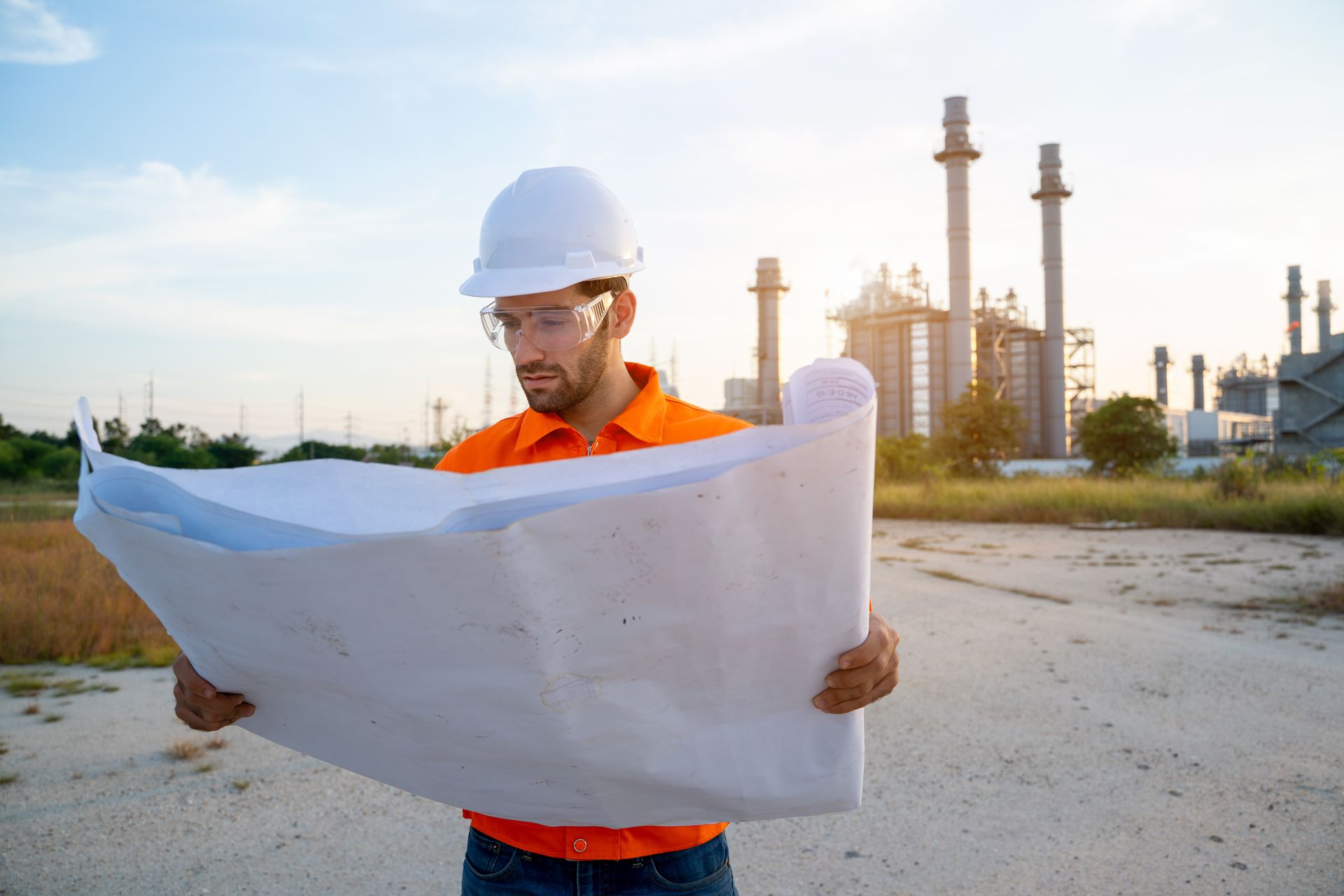 Un ingénieur, portant un casque et des lunettes de sécurité, examine des plans près d'une installation industrielle.