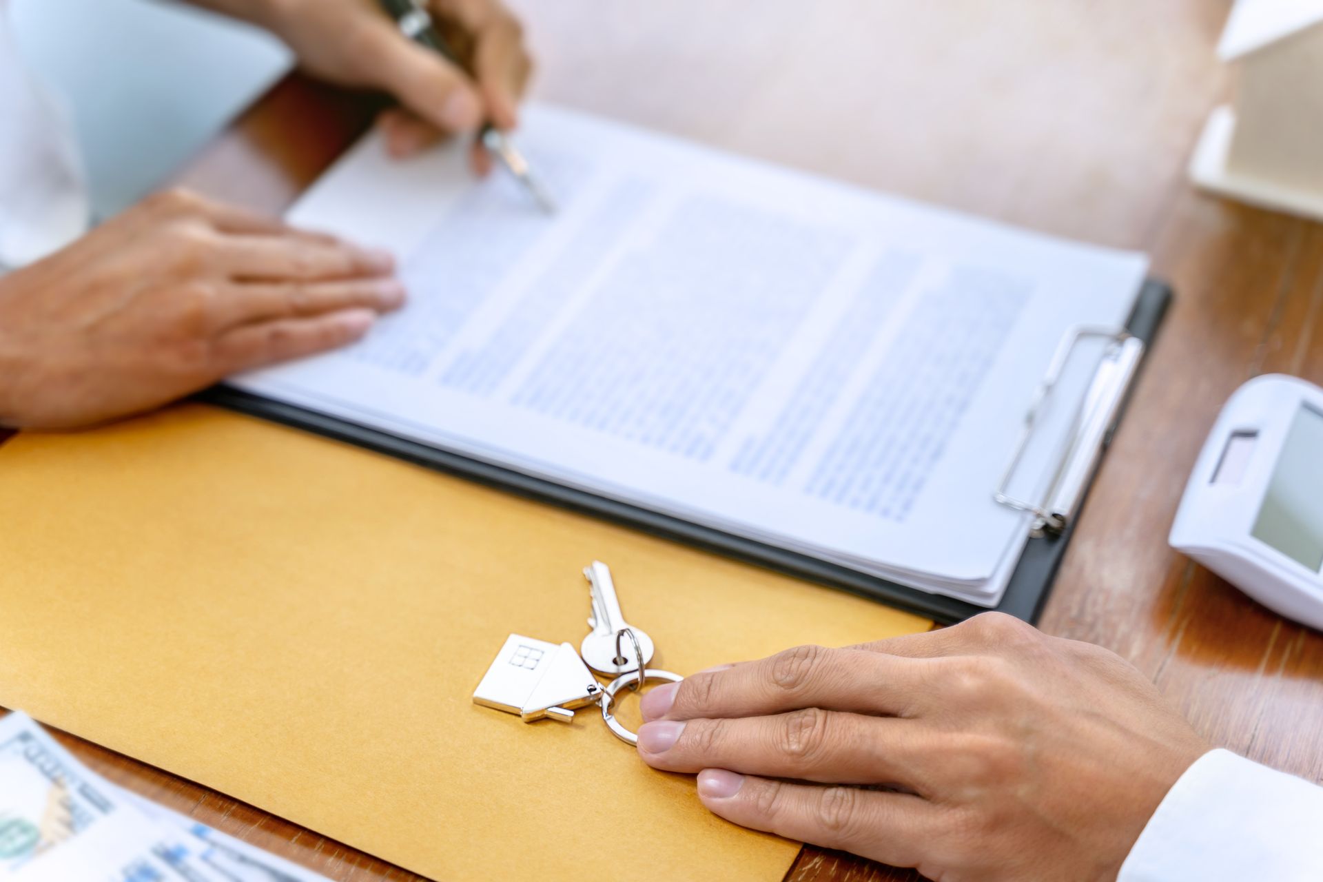 Des mains signant un document, des clés ornées d'un pendentif en forme de maison sur une table et de l'argent liquide dans une enveloppe en papier kraft.