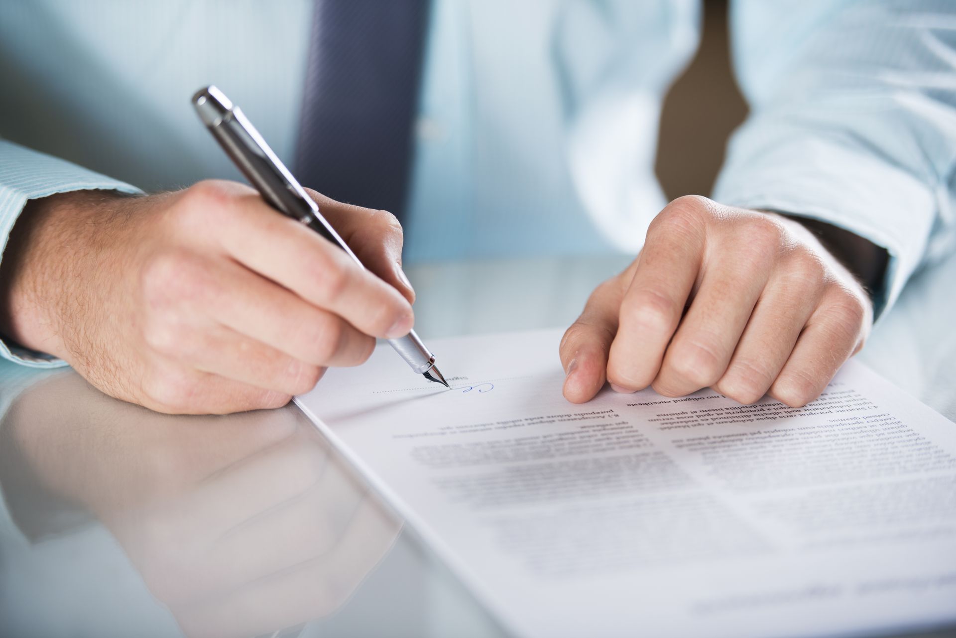 Une personne en chemise bleue et cravate signe un document avec un stylo argenté sur une table blanche.
