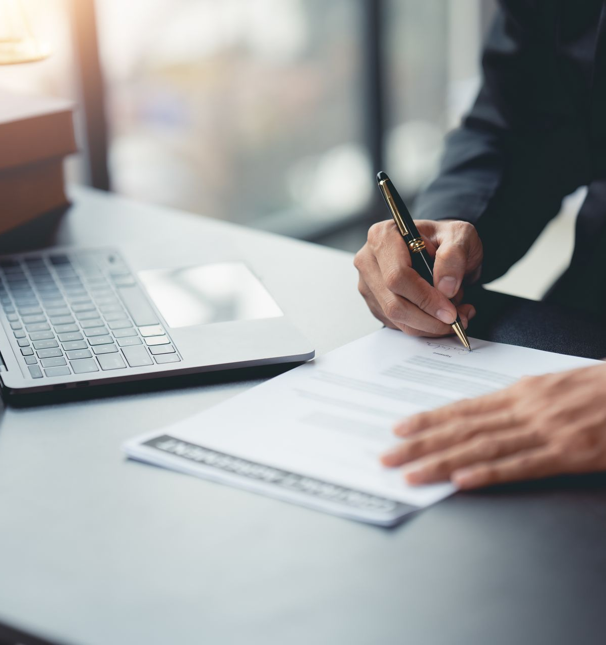 Une personne en costume écrit sur du papier avec un stylo à un bureau, son ordinateur portable à proximité.