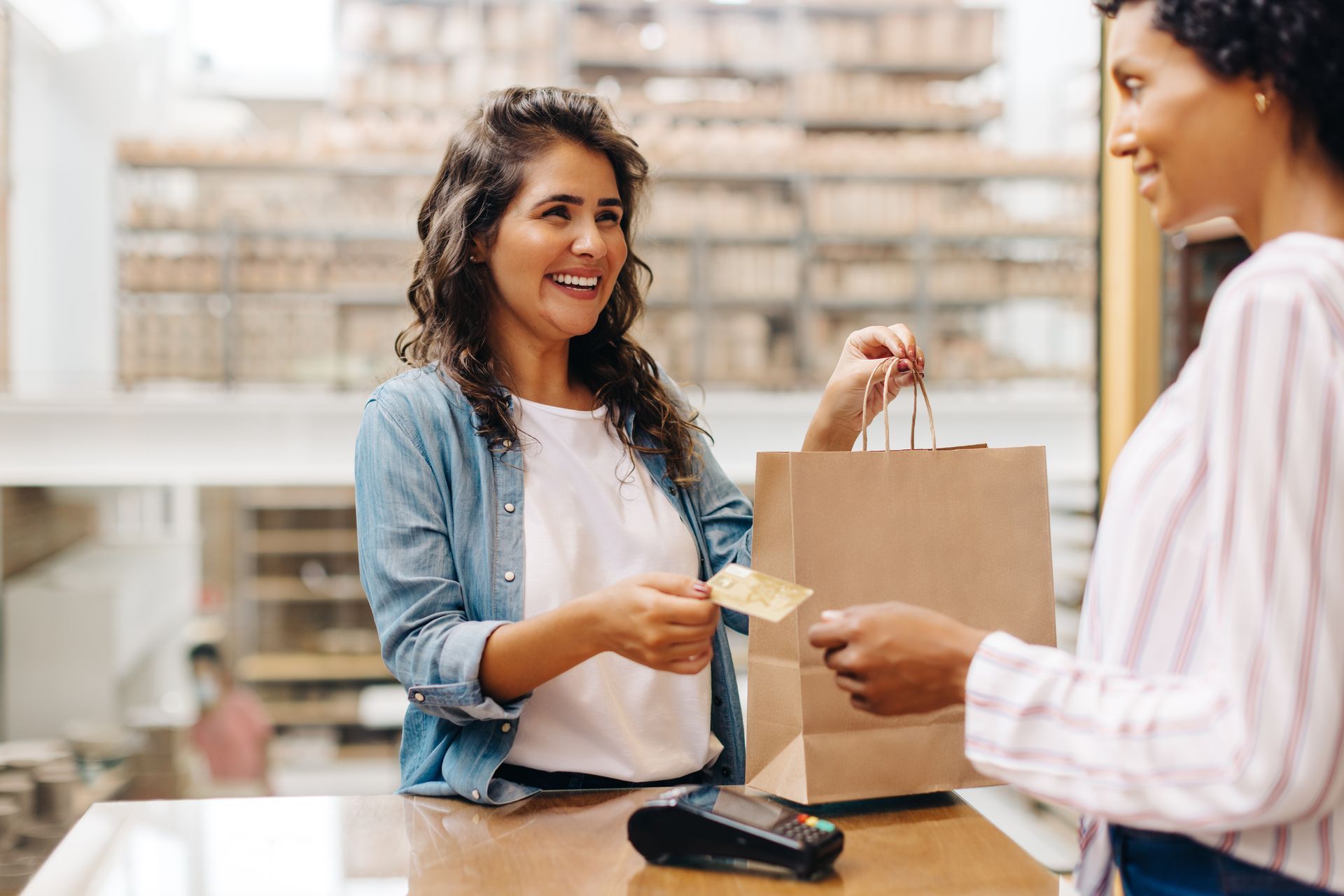 Une femme paie par carte bancaire à la caisse d'un magasin ; une autre femme lui tend un sac de courses.