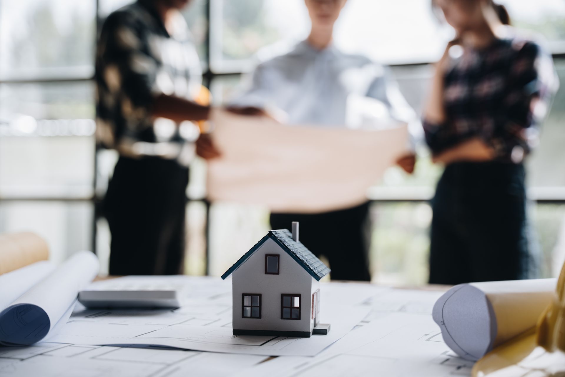 Maison miniature sur une table à plans, avec trois personnes examinant les plans en arrière-plan.